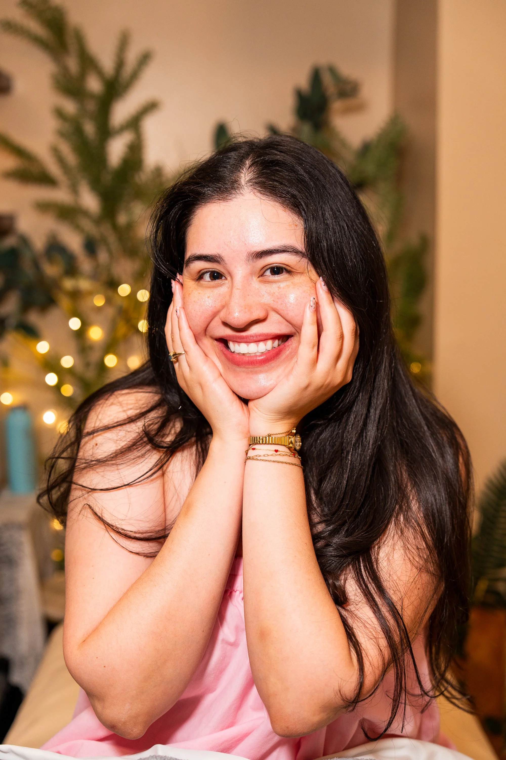 A woman with long dark hair smiling and resting her face in her hands, wearing a pink satin dress, in front of a blurred background with Christmas lights and green plants.