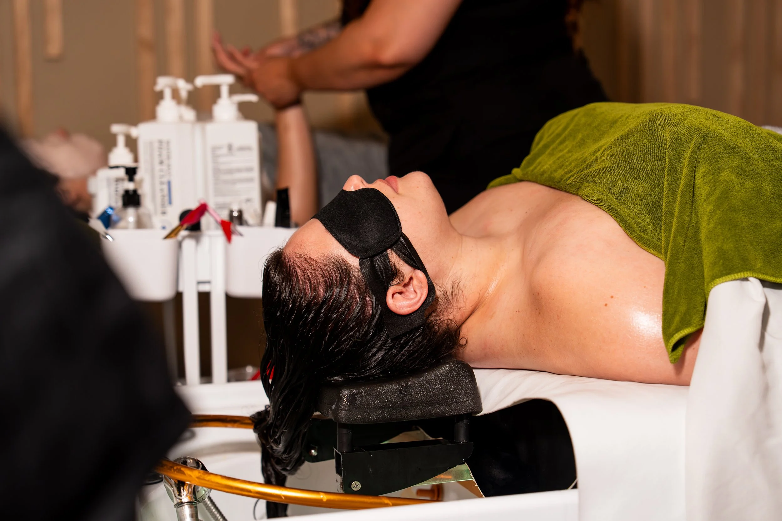 A woman lying on a medical table with a black sleep mask, receiving cosmetic or skincare treatment, with various bottles and medical equipment nearby.