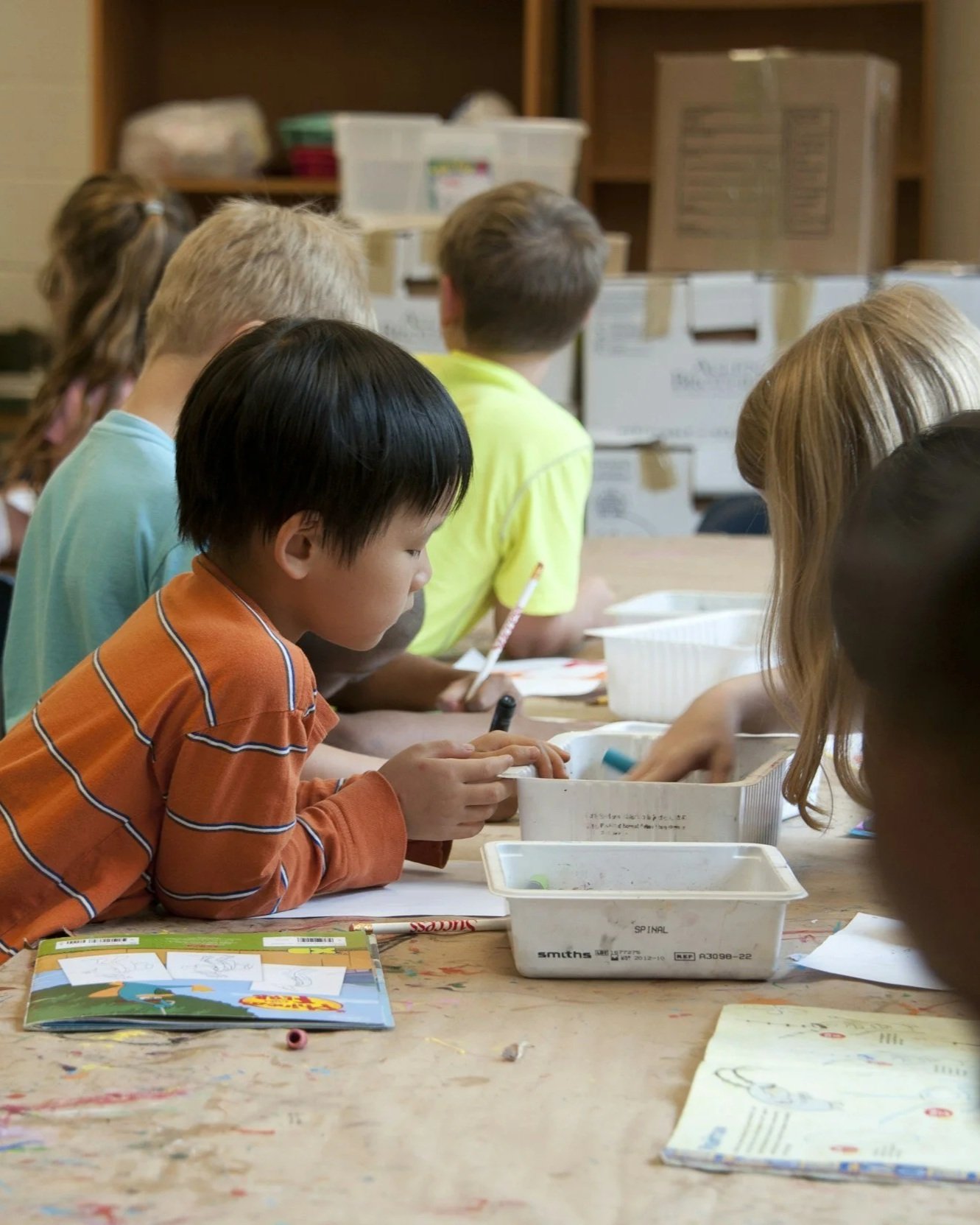 Children engaged in arts and crafts at a table with drawing supplies, books, and bins in a classroom setting.