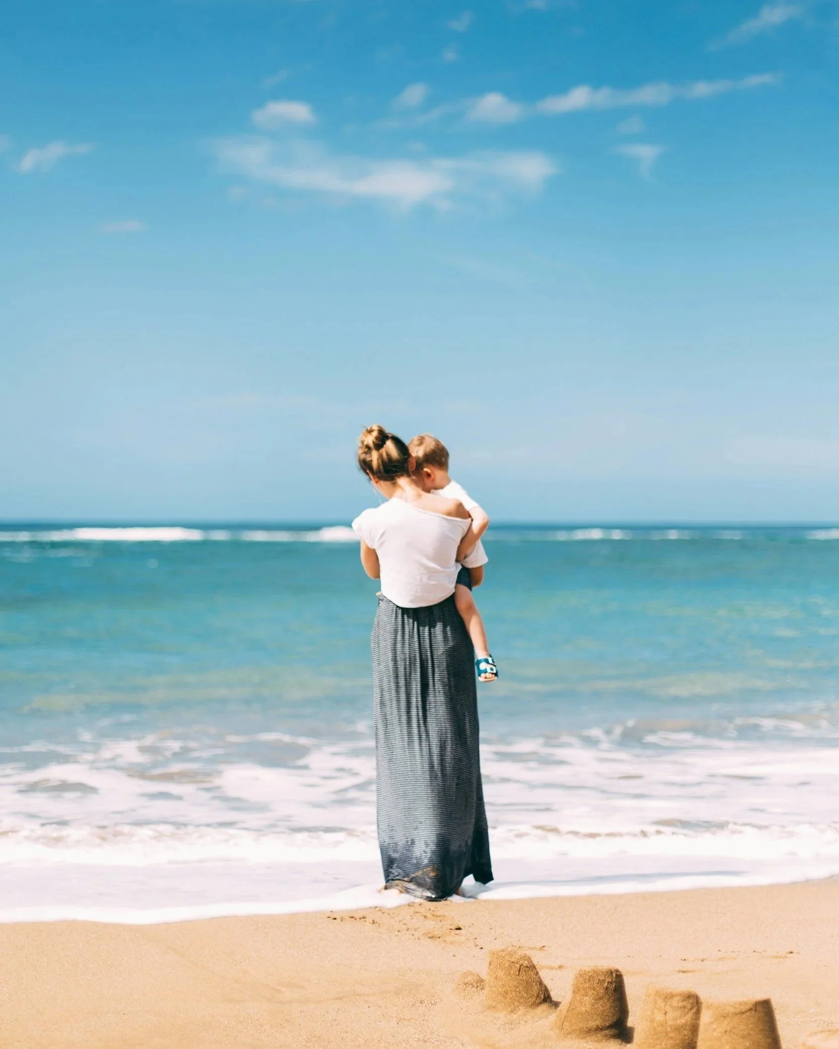 A woman standing on the beach holding a young child, with sandcastles in the foreground, the ocean and blue sky in the background.