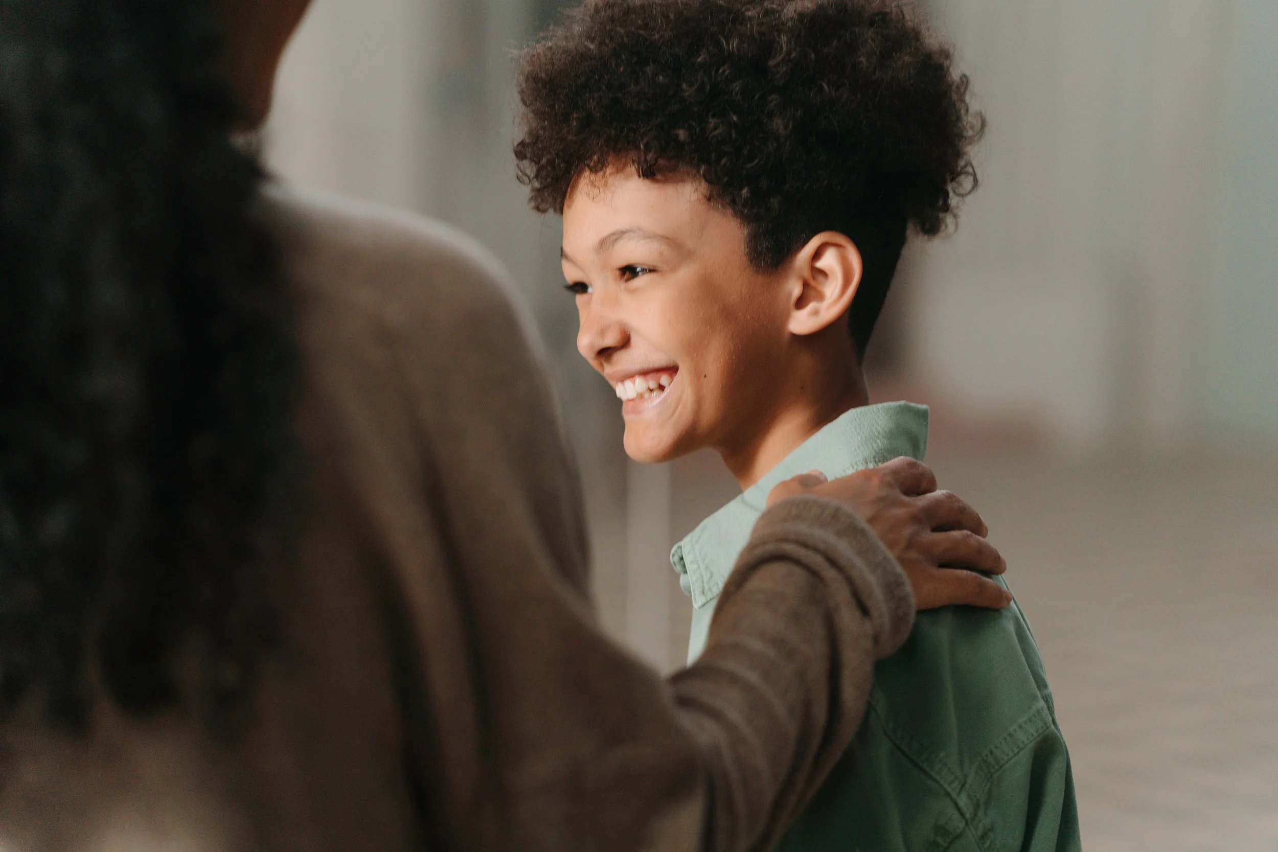 A young boy with curly hair smiling while being comforted or hugged by an adult.