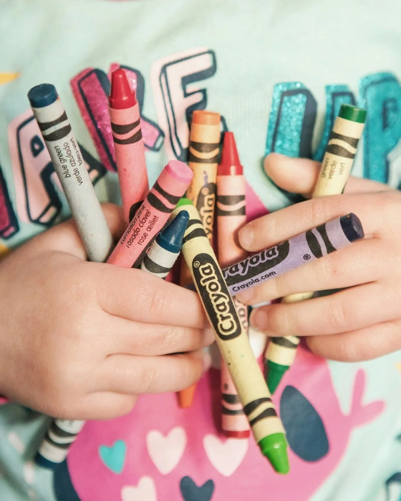 Child holding multiple Crayola crayons in front of a colorful background with the word 'LOVE' and hearts.