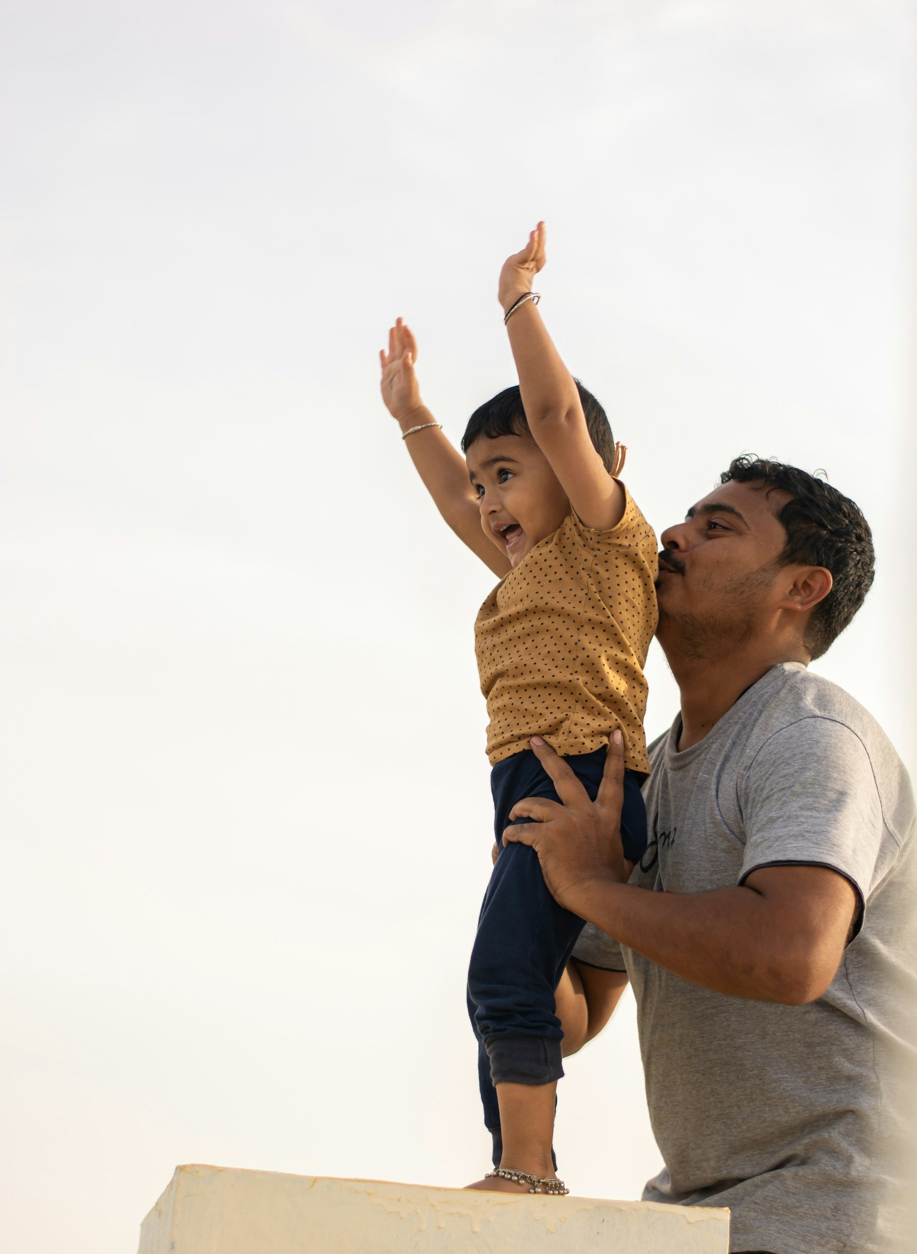 A young boy wearing a yellow polka dot shirt and dark blue pants is sitting on an adult man's shoulders. The boy has his arms raised in the air, smiling with excitement. The man, dressed in a gray t-shirt, is holding the boy securely while looking up at him. The background features a clear sky.