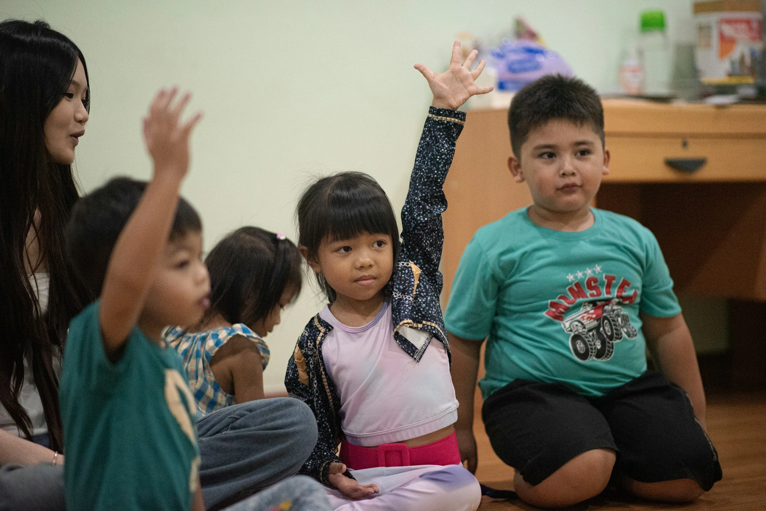 Children seated on the floor during a classroom activity, with one girl raising her hand.