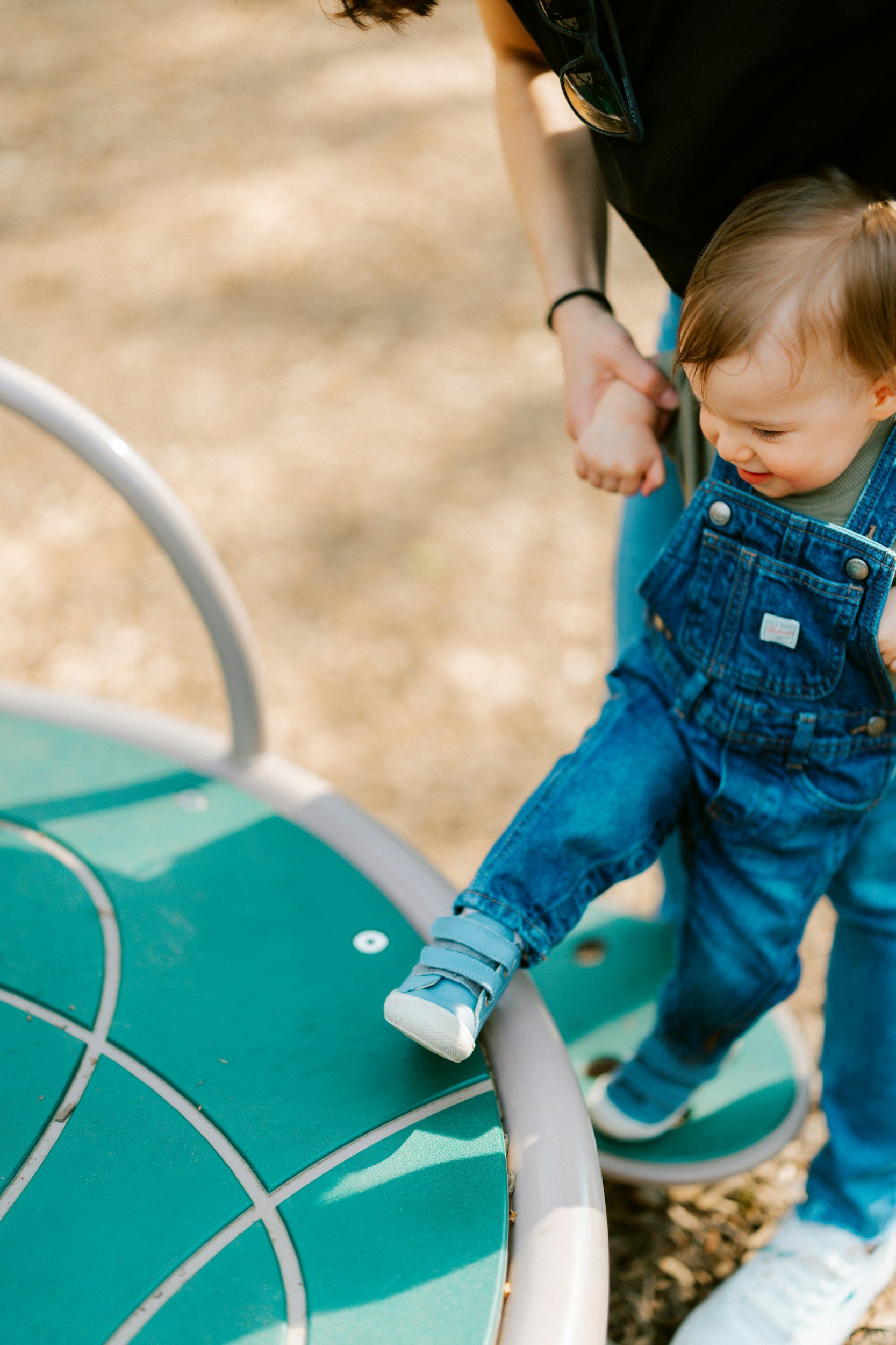 A young child with a denim jacket and gray sneakers holding an adult's hand near a green and white playground structure outdoors.