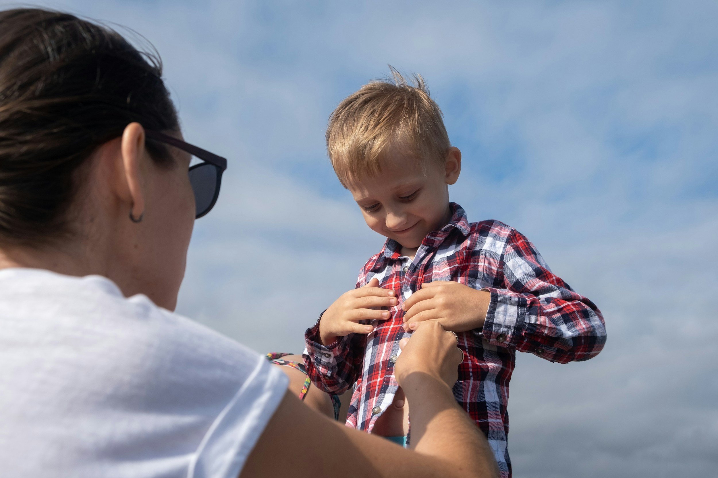 A child wearing a plaid shirt being helped by adults to put on a red plaid shirt outdoors, with a blue sky and clouds in the background.