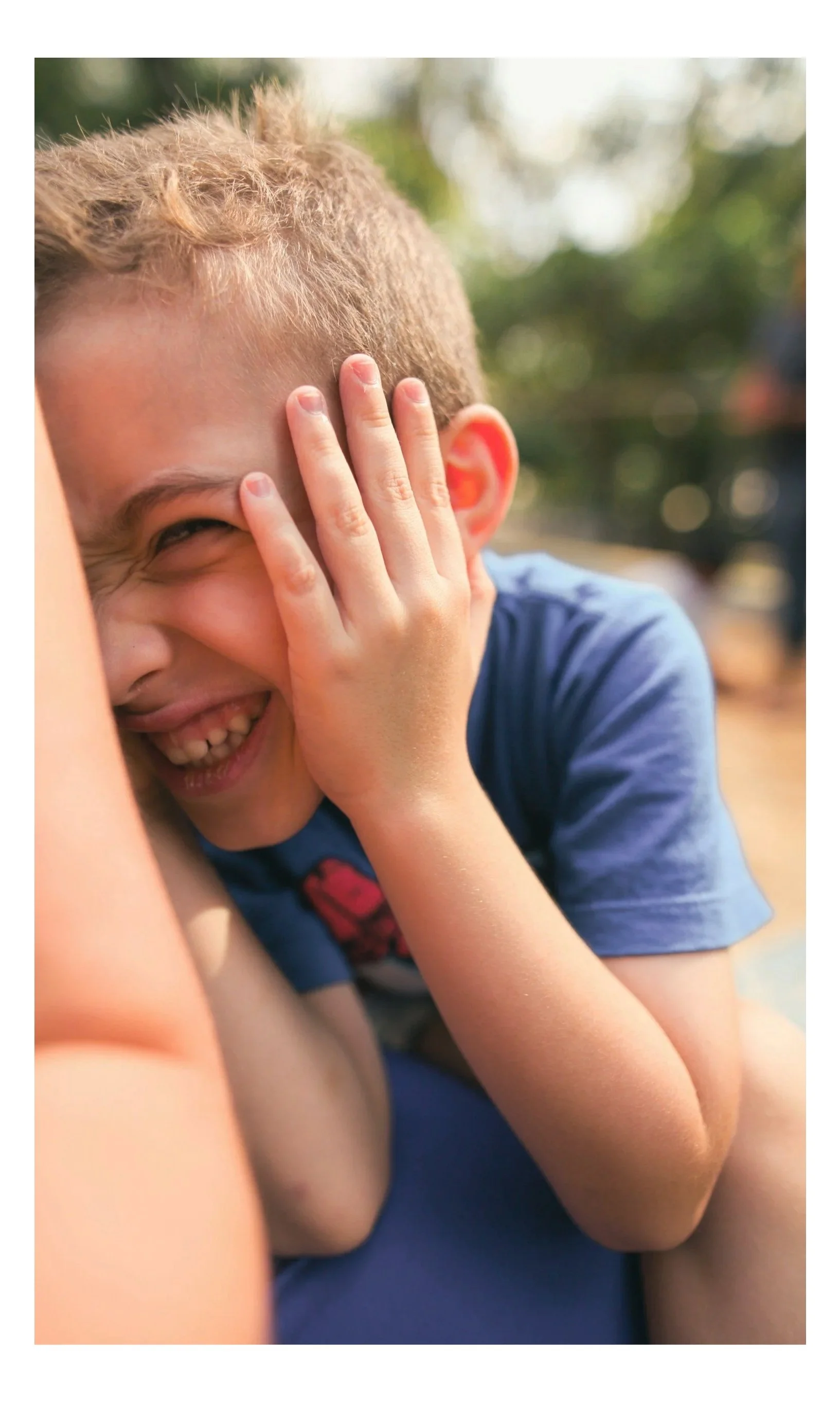A young boy laughing and covering his face with his hand, showing a joyful expression. He's wearing a blue shirt and appears to be outdoors with blurred trees in the background.