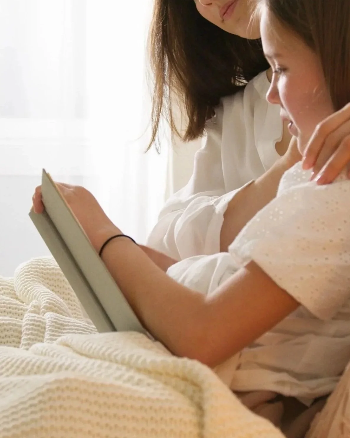 A woman and a girl sitting on a bed looking at a phone together, with the woman leaning over the girl, in a bright room.