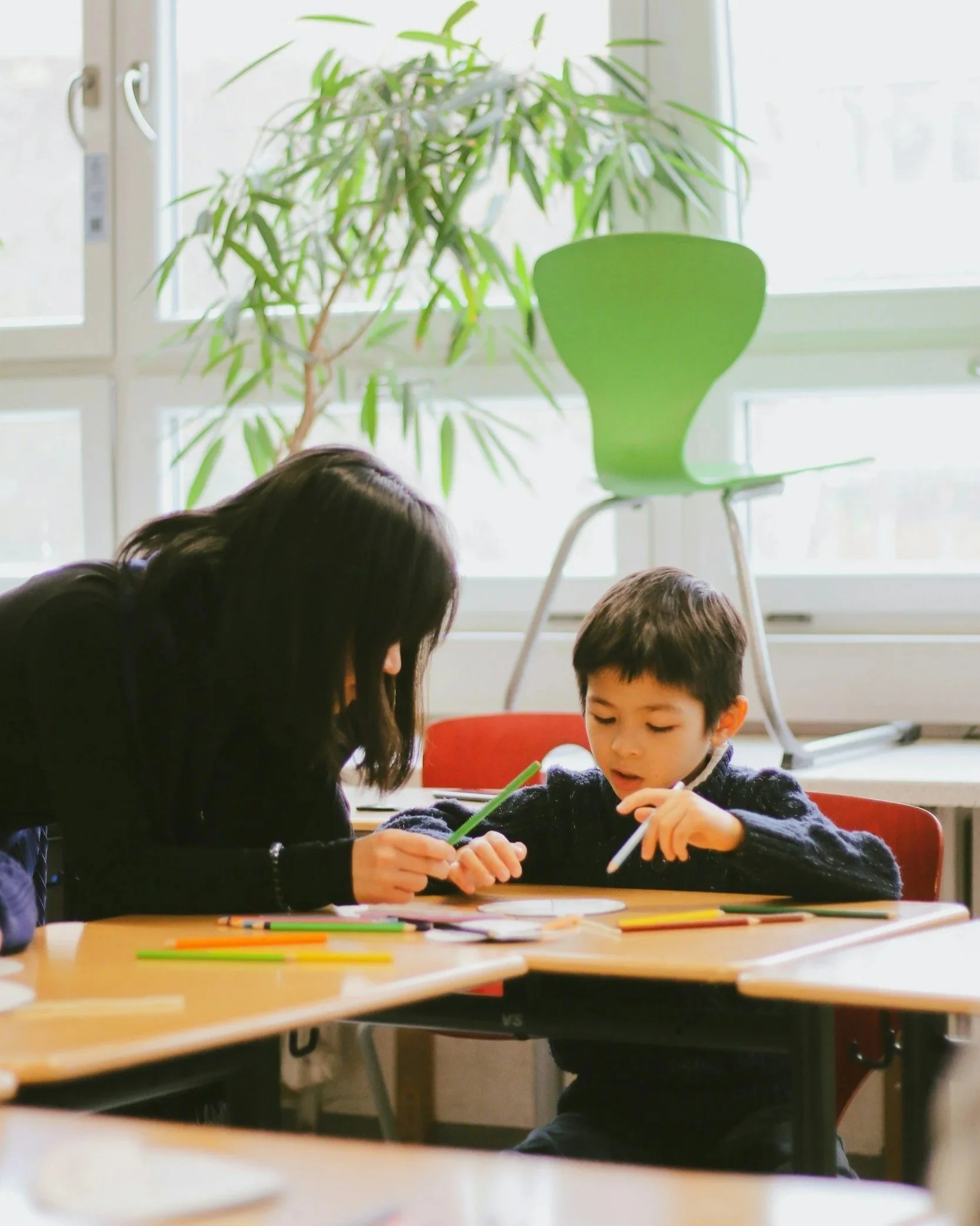 Kids sitting at a table, drawing with colored pencils, near a window with a green plant and a lime green chair.