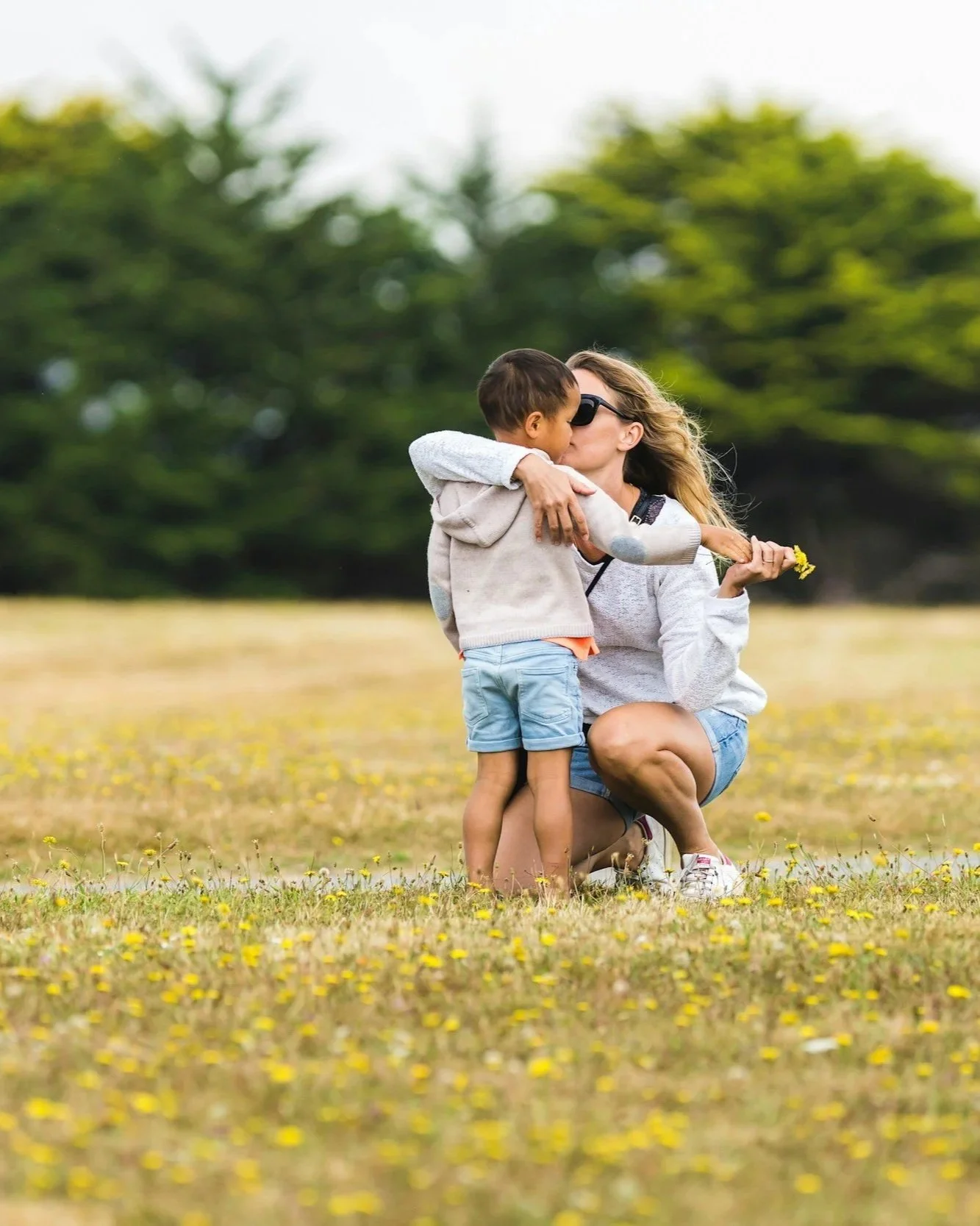 A woman kneeling on the grass with yellow flowers, hugging a young boy and kissing him on the cheek outdoors.