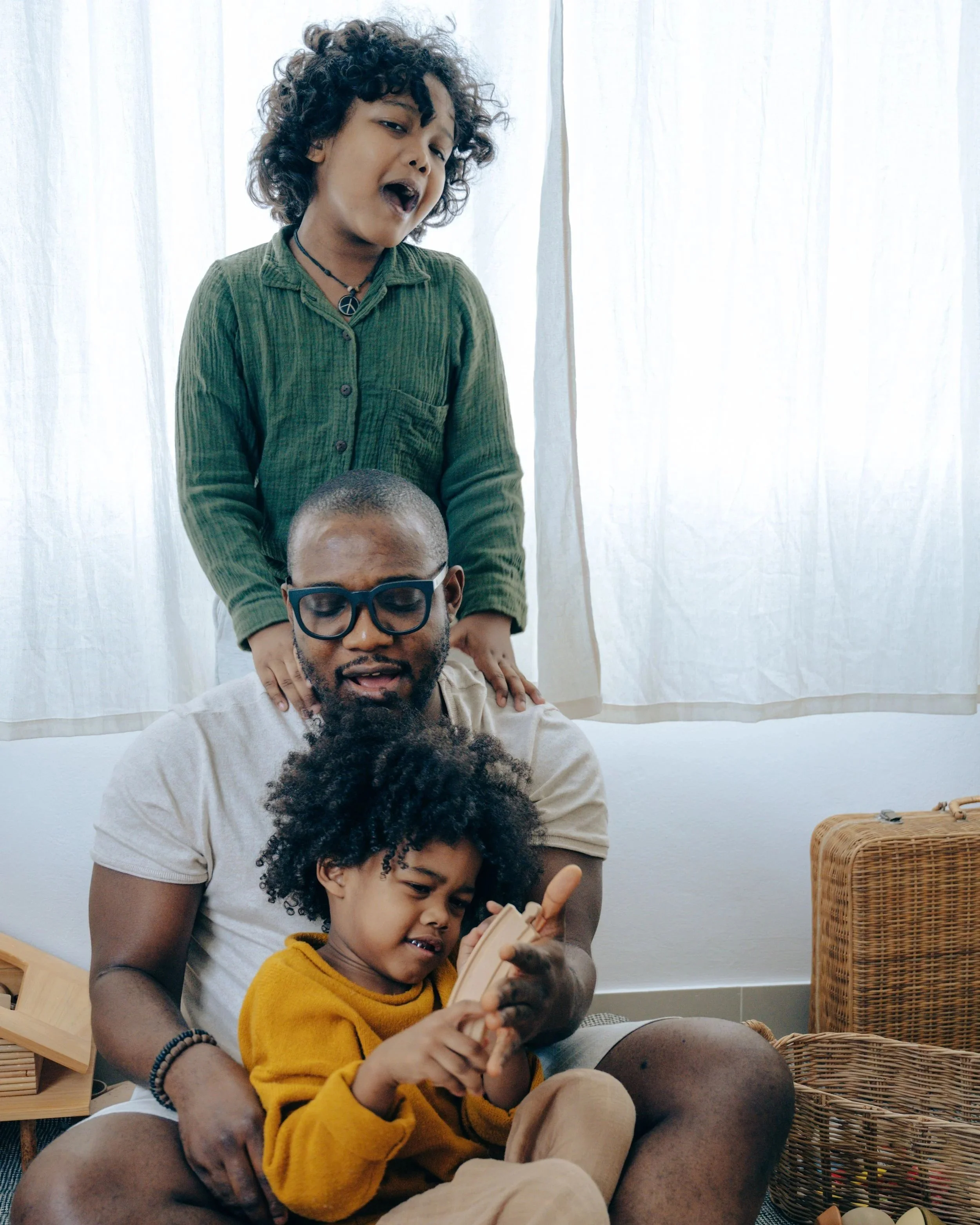 A woman with curly hair and a peace necklace standing behind a seated man with glasses and a beard, who is in a meditative pose. A young girl with curly hair in a yellow sweater is sitting on the man's lap, looking at a device. The background includes white curtains and wicker baskets.