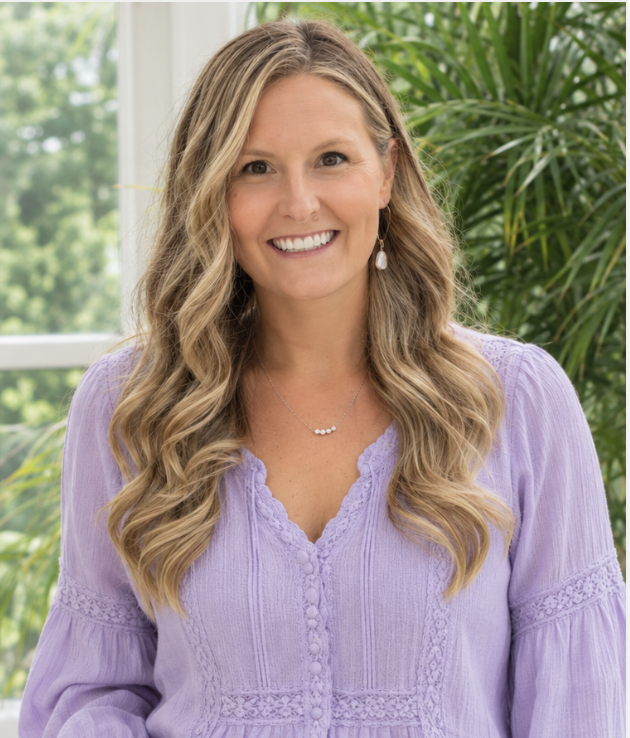 A smiling woman with long wavy blonde hair wearing a lavender blouse with lace details, standing indoors near a window and large green plants.