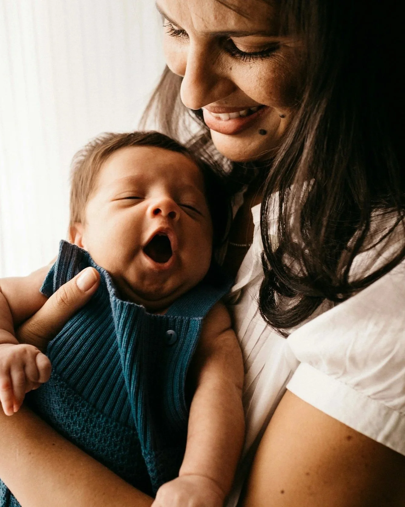 A woman with long dark hair holding a yawning baby with closed eyes, wearing a blue sleeveless shirt, close to a white wall, captured in a warm, intimate moment.