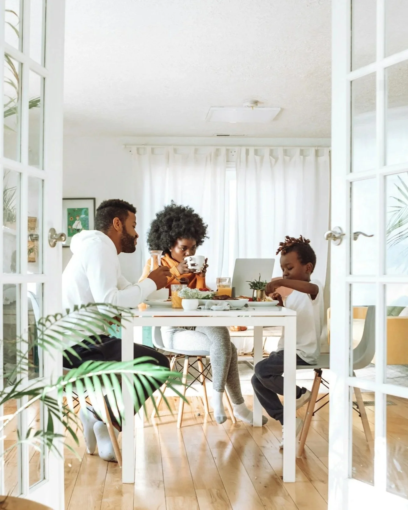 A family of three, two adults and a child, sitting at a dining table enjoying breakfast in a bright, cozy kitchen with white curtains, plants, and artwork on the walls.