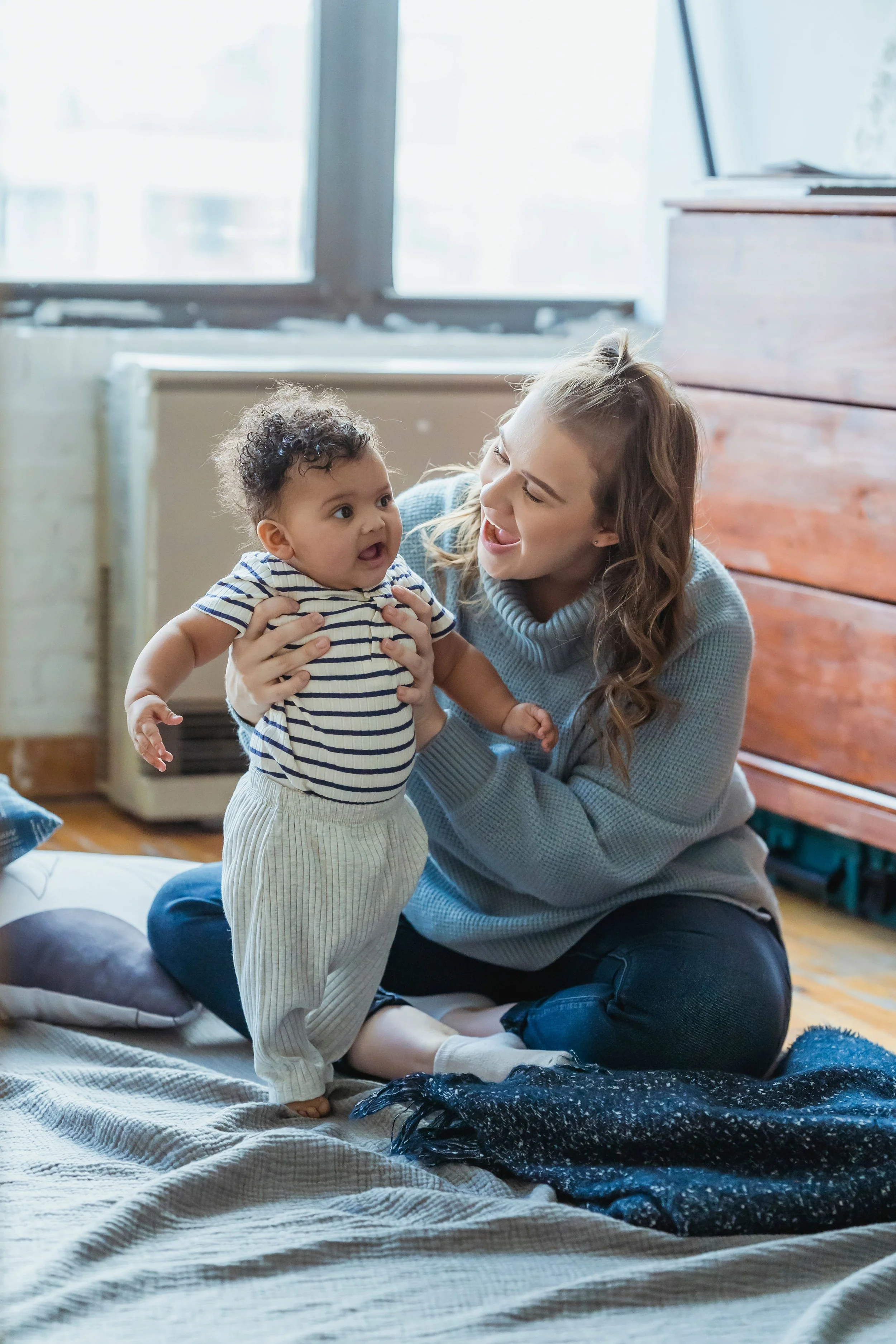 A woman and a young child playing together on a blanket inside a room with large windows, a dresser, and a radiator.