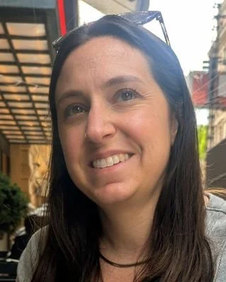 A woman with dark hair, wearing glasses on her head, smiling in an outdoor setting.