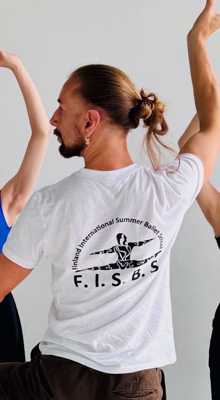 Person with long hair tied in a bun, wearing a white T-shirt with a logo of Finland International Summer Ballet Festival F.I.S.B. and participating in a ballet pose with arms raised.