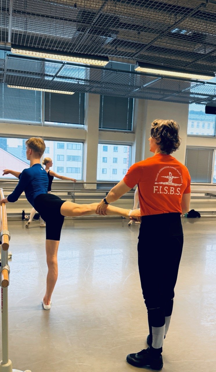 A ballet instructor assisting a young dancer with a leg stretch at a dance studio. The instructor wears an orange shirt with 'F.I.S.B.S.' on the back. The studio has large windows and ballet barres.