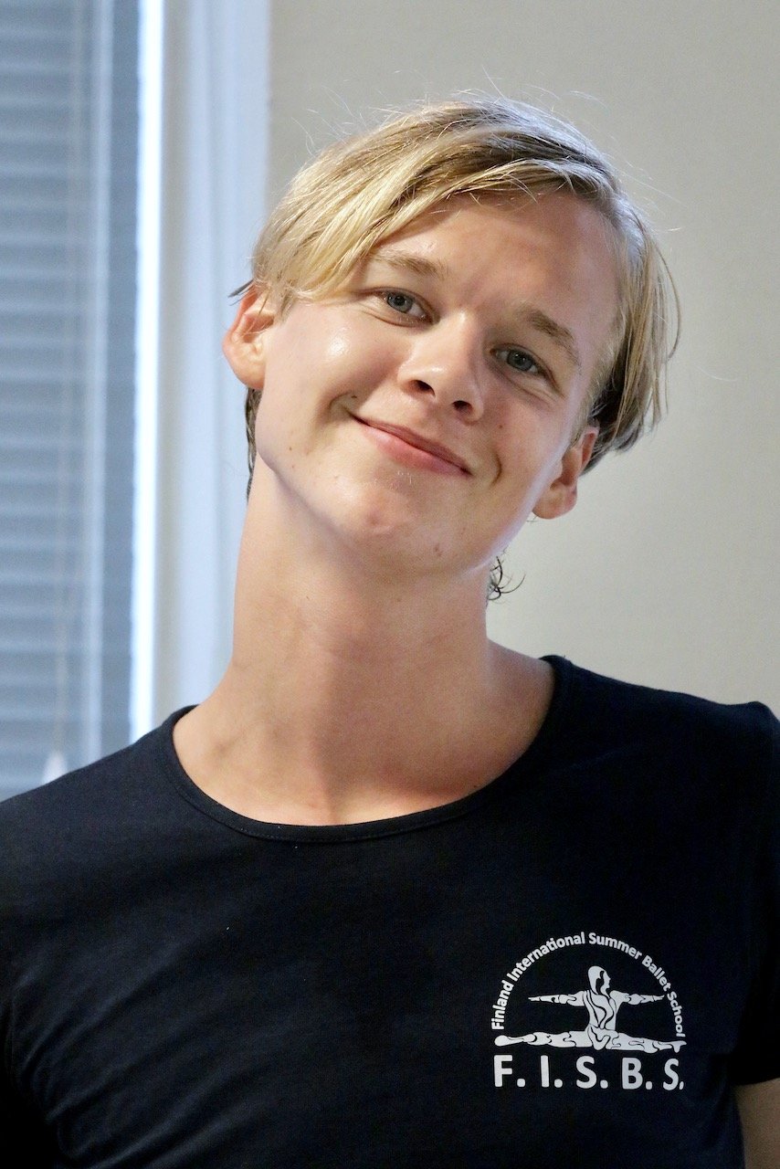 A young person with short blonde hair smiling, wearing a black t-shirt with a logo for the Finland International Summer Ballet School.