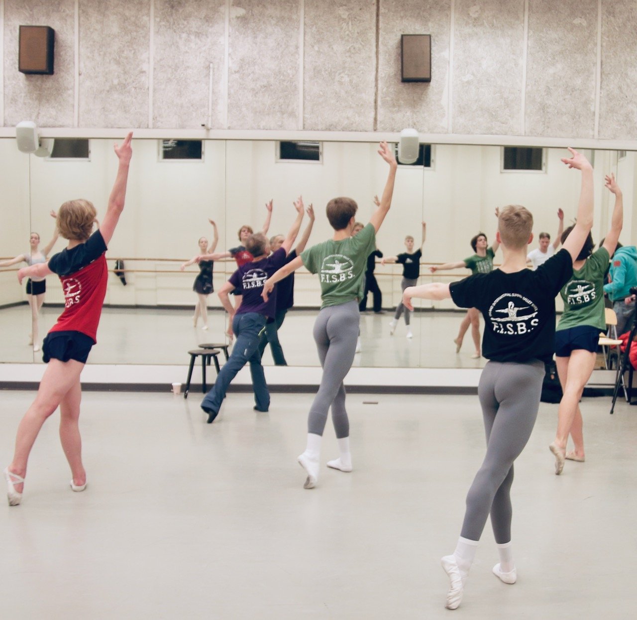 Ballet dancers practicing in a dance studio with a mirrored wall, some dancers wearing colorful shirts with the letters "F.I.S.B.S." and gray leggings, performing ballet movements.