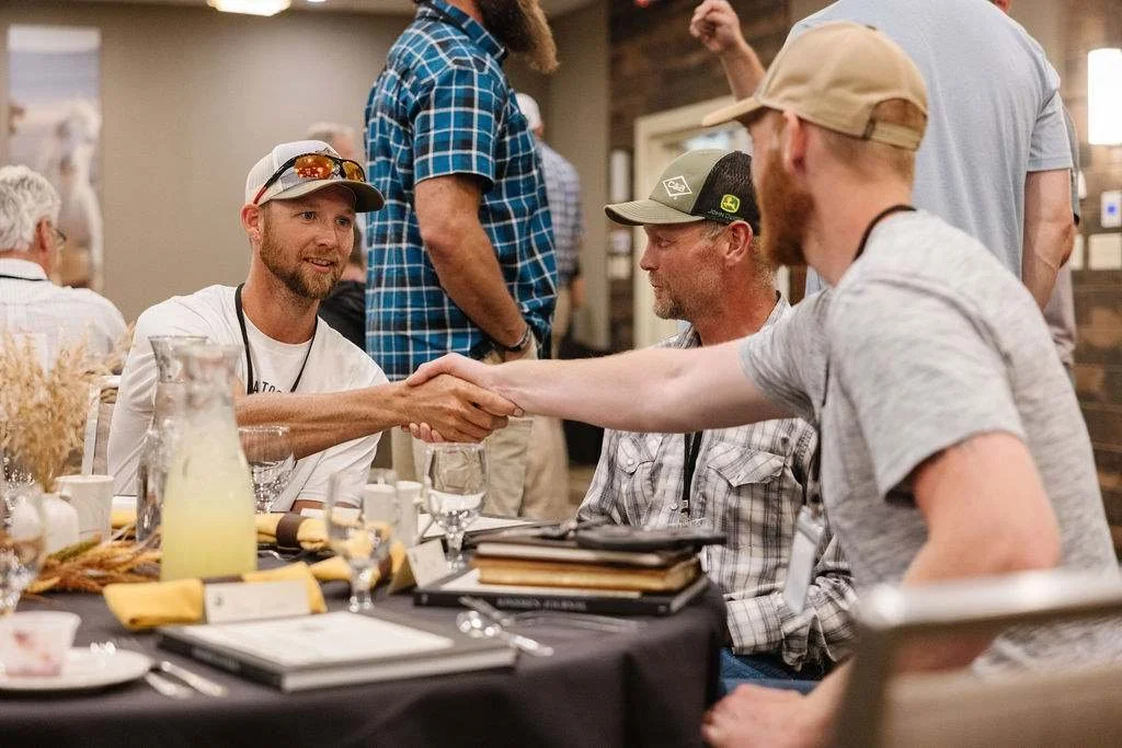 Two men shaking hands at a table during a social gathering, with others sitting around and standing nearby, in a restaurant or cafe setting.