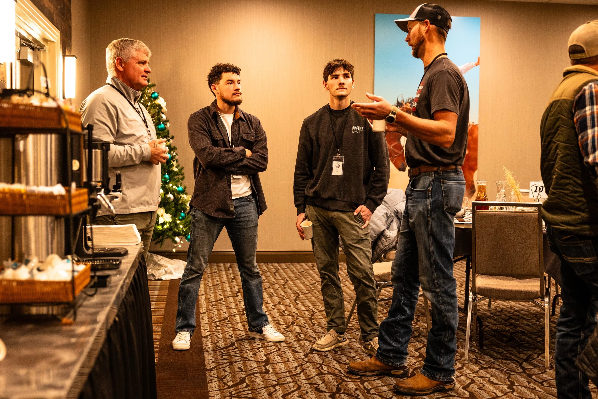 Four men are standing and talking in a conference room, with a Christmas tree in the background. The men are engaged in conversation, with some holding cups, surrounded by tables with food and drinks.