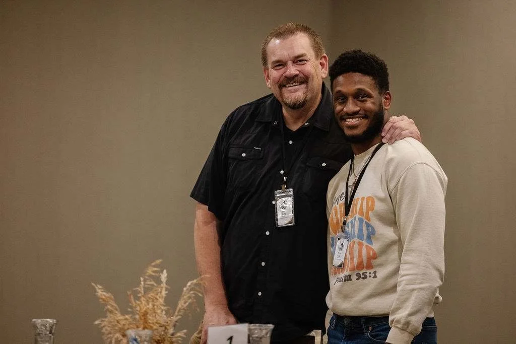 Two men smiling, standing close together, with one man having his hand on the other's shoulder. They are indoors, with a plain wall in the background. The man on the left has a beard and is wearing a black shirt with ID badge, and the man on the right has a beard and is wearing a beige sweatshirt with colorful text and an ID badge. There are some decorative dried plants and a small sign in front of them.