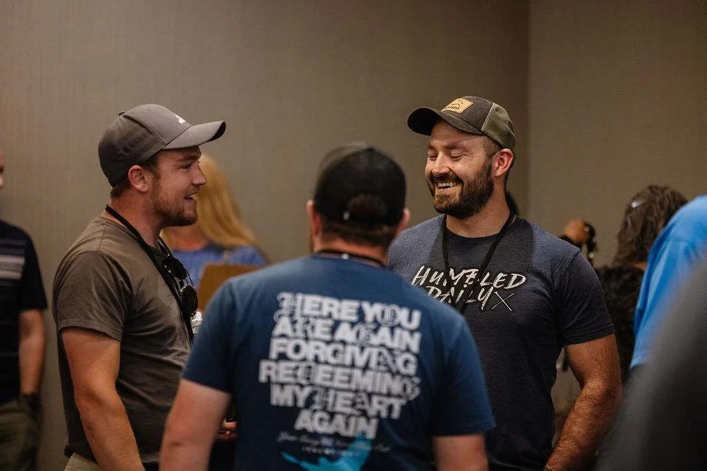 Three men engaged in conversation, smiling, in a crowded indoor setting.