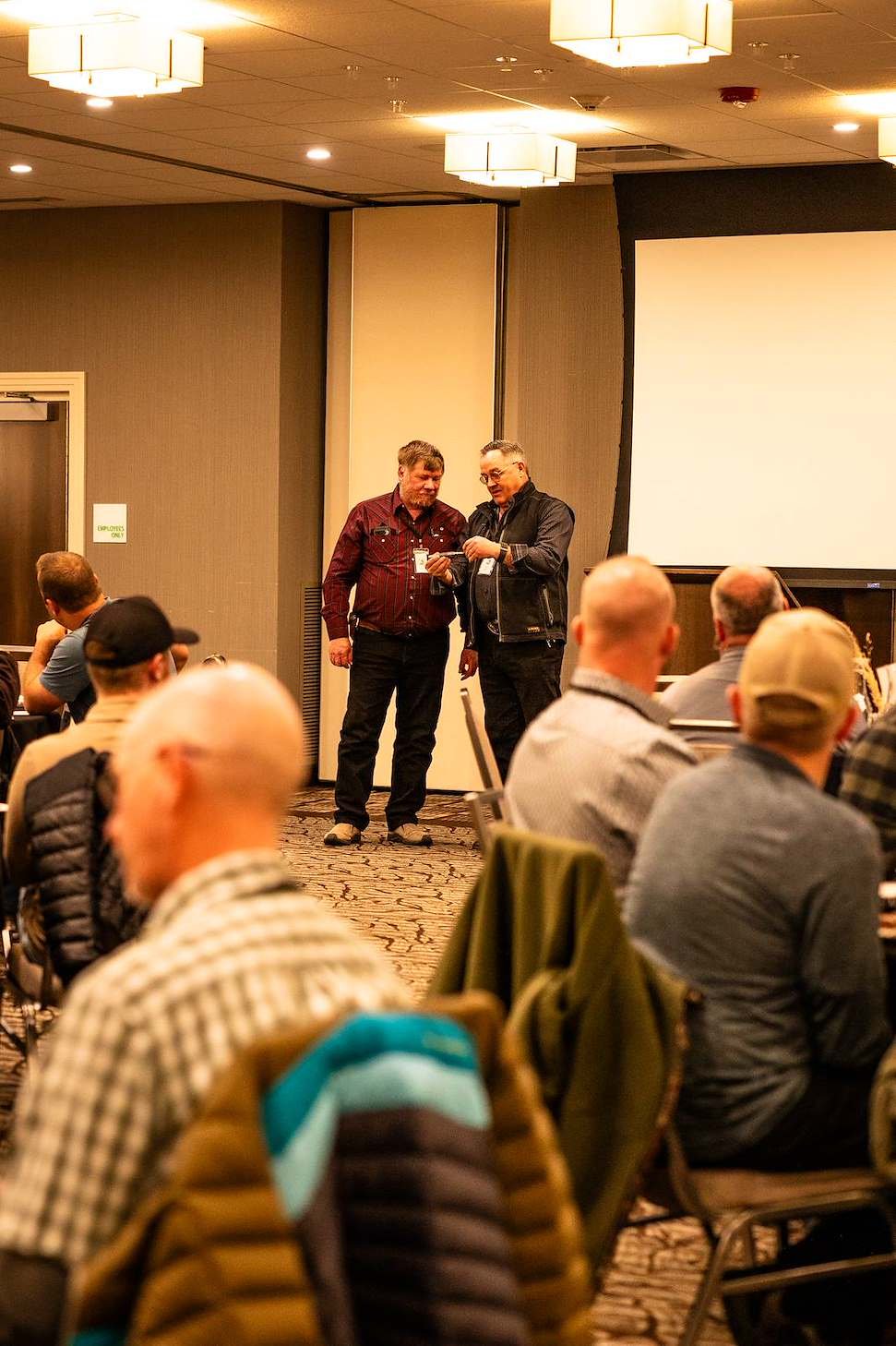 Two men stand near the front of a conference room, speaking to each other, while an audience listens. The room has ceiling lights, a large blank projection screen, and an elevator with a sign.