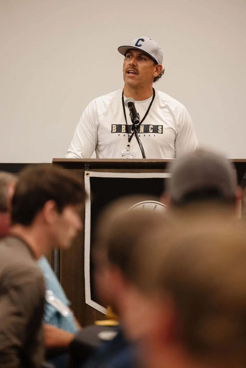 Man wearing baseball cap and white sports shirt speaking at a podium during a meeting with an audience.