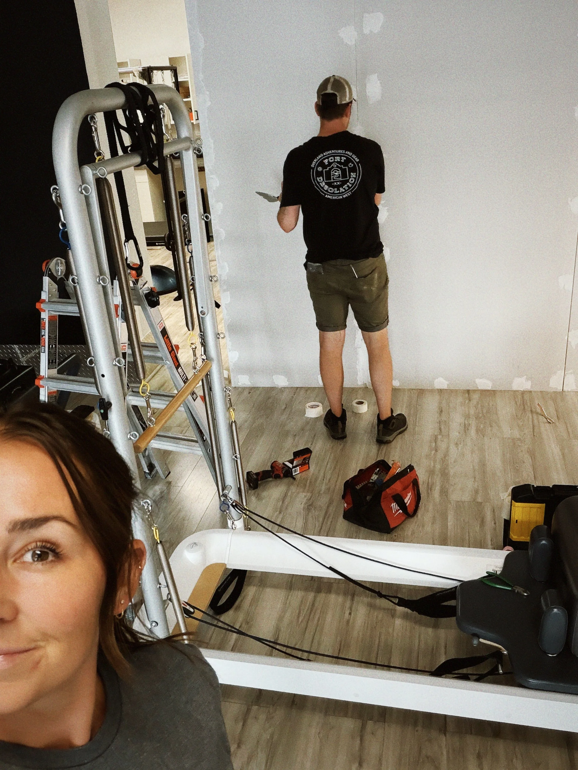 A person with glasses and brown hair taking a selfie in front of a mirror under construction, with drywall patches visible. In the background, another person is working on the wall, with tools and supplies scattered on the floor.