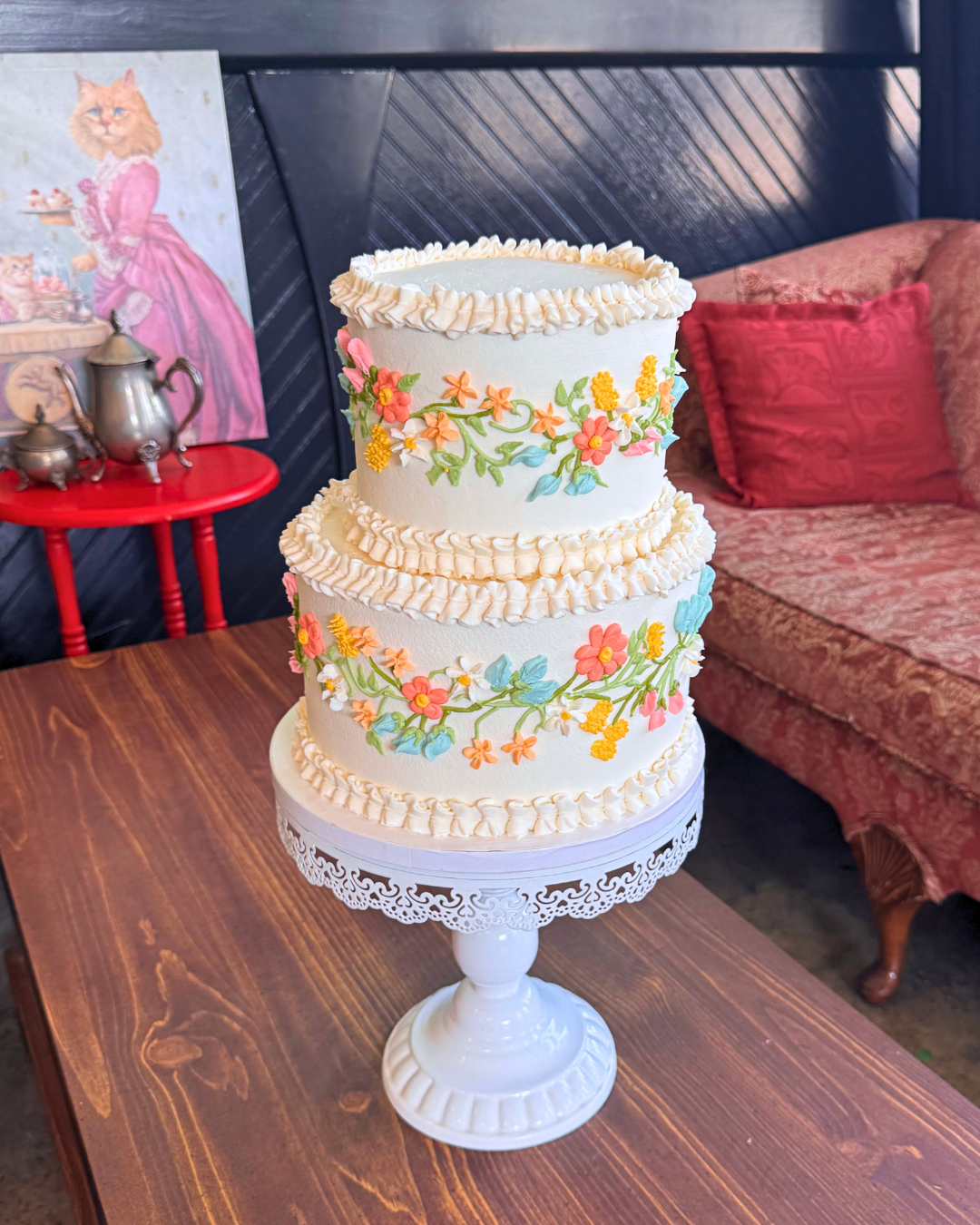 A three-tiered white wedding cake decorated with pastel-colored floral icing, placed on a white cake stand on a wooden table.