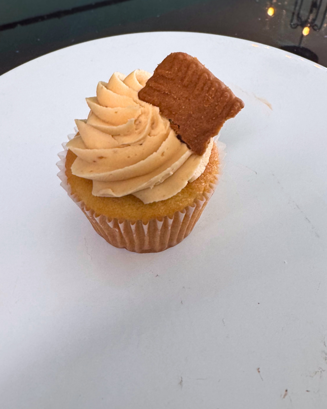 Cupcake with beige frosting, topped with a small chocolate cookie, on a white surface.