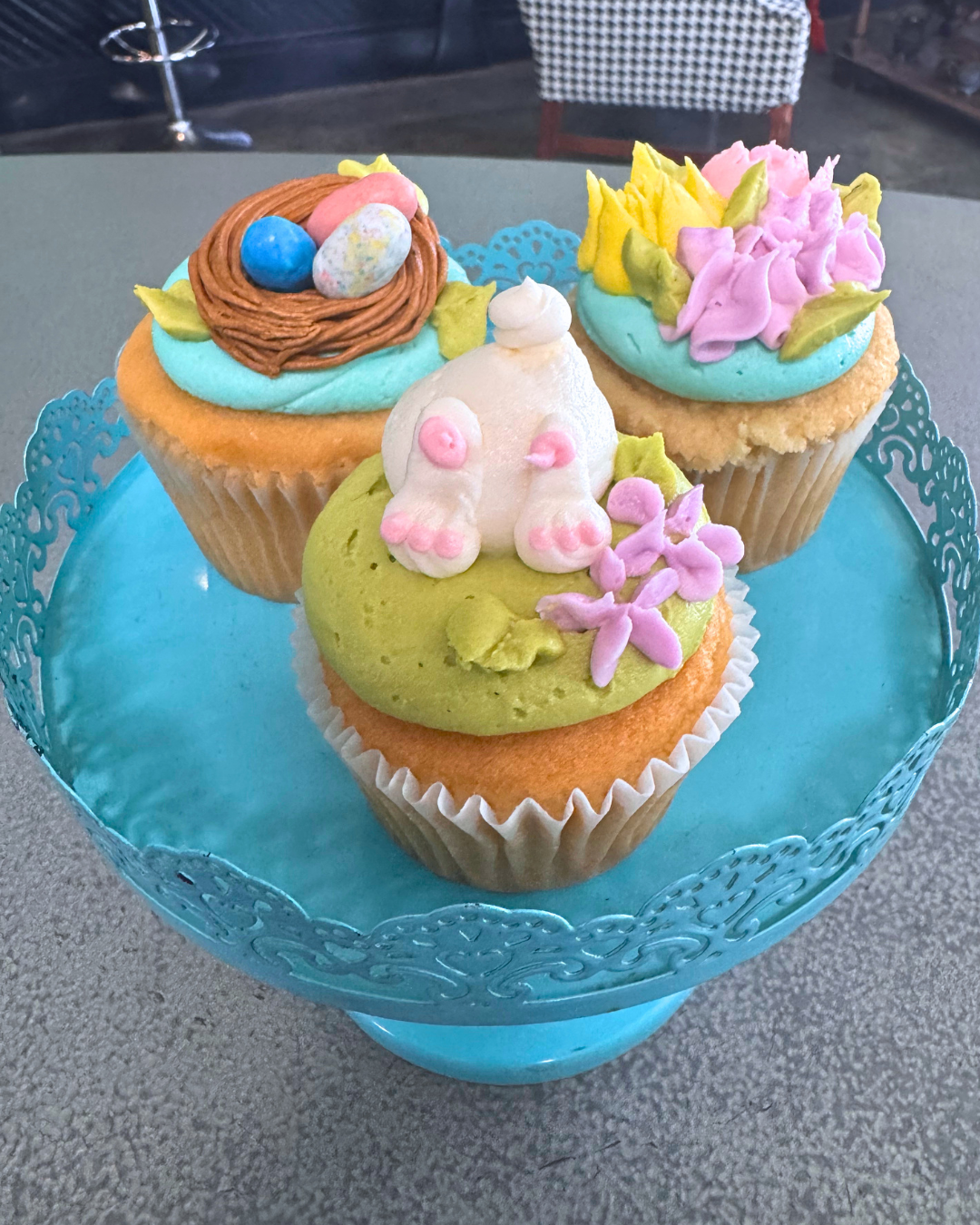 Three decorated cupcakes in a blue lace-patterned cupcake stand, each with pastel-colored frosting and decorative toppers, including a nest with eggs, a bunny, and flowers.
