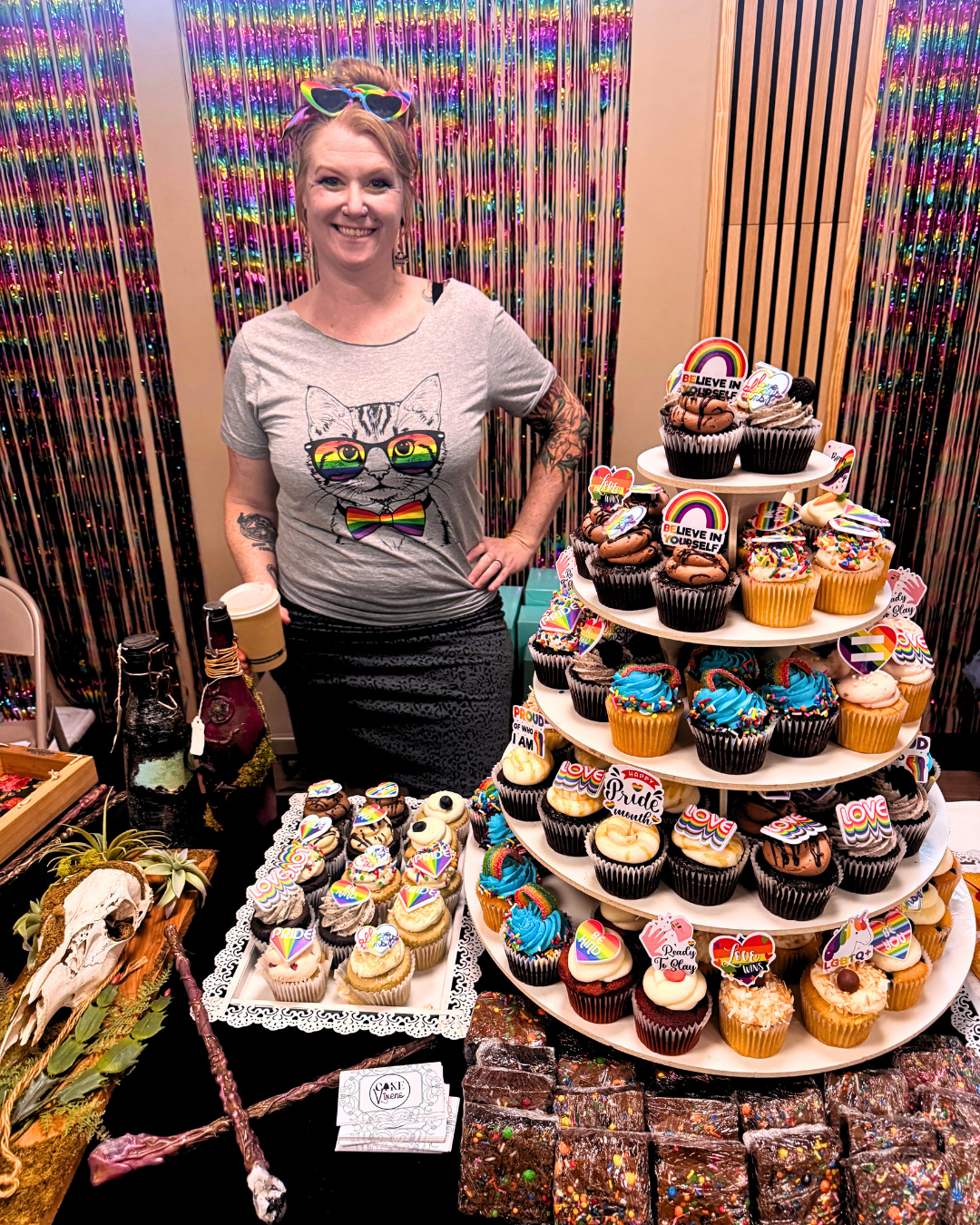 A woman standing behind a display of rainbow-themed cupcakes with colorful decorations, including rainbow toppers and pride symbols, at a celebration event with a multicolored shiny backdrop.