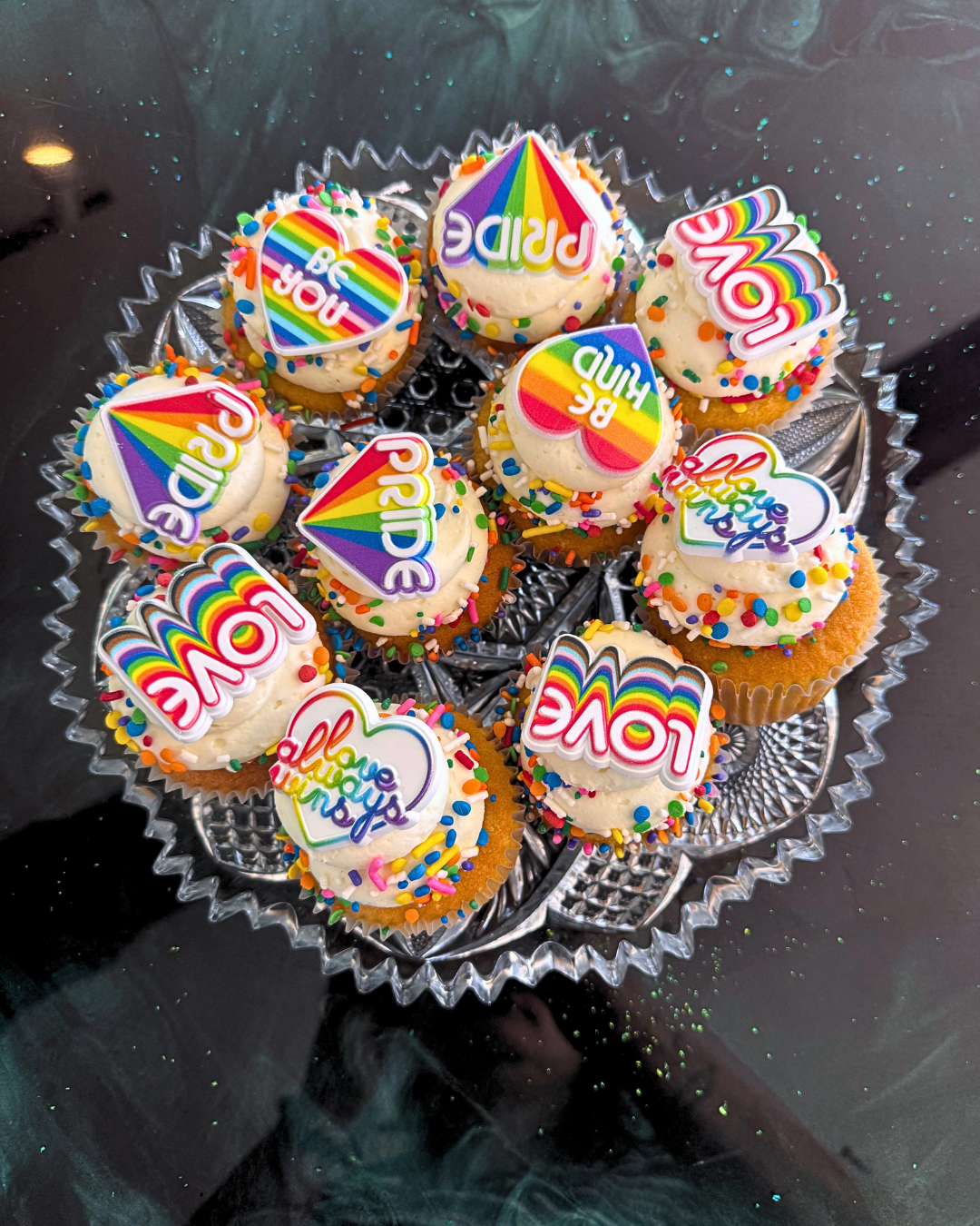 Cupcakes decorated with colorful rainbow-themed 'Love You' and 'Be Happy' toppers, topped with sprinkles, arranged on a silver tray.
