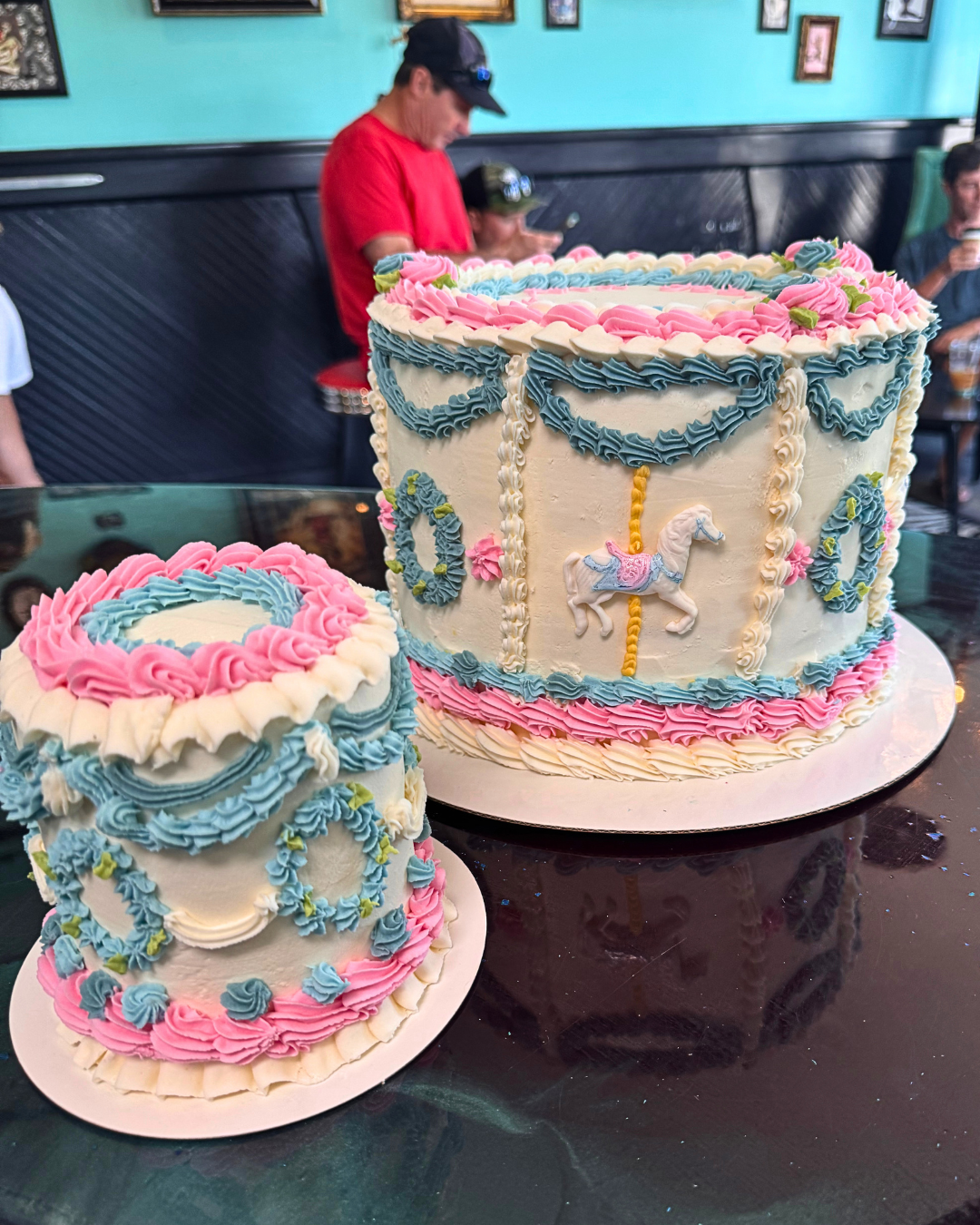A birthday cake decorated with blue, pink, and white icing featuring a carousel horse design, located on a black table with a smaller matching cake in front of it, in a room with teal walls and framed pictures. People in the background are engaging in conversation and using their phones.