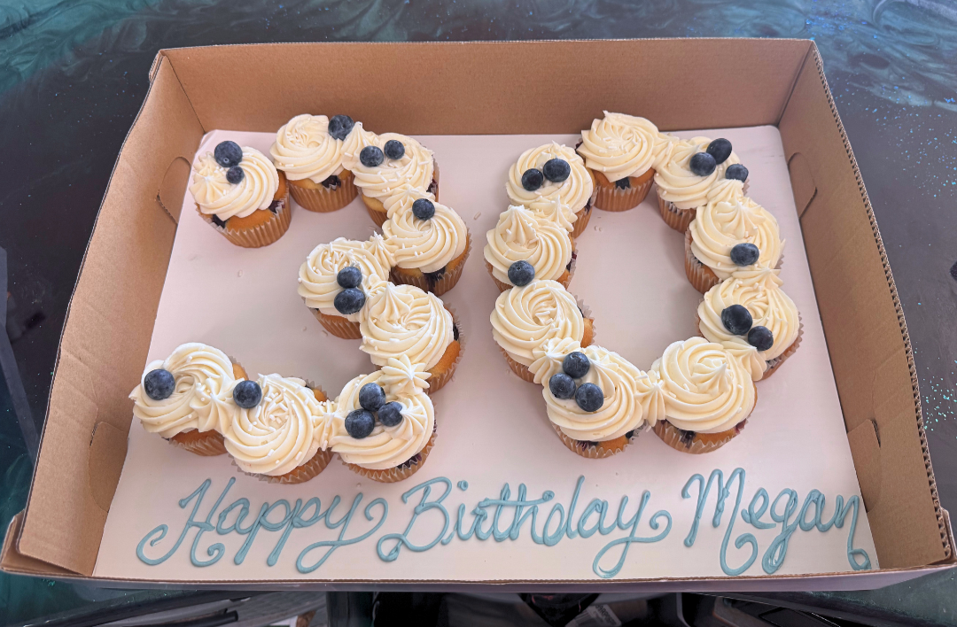 Cupcakes arranged in the shape of the number 30, decorated with white frosting and blueberries, in a bakery box. The message 'Happy Birthday Megan' is written on the cake's surface.