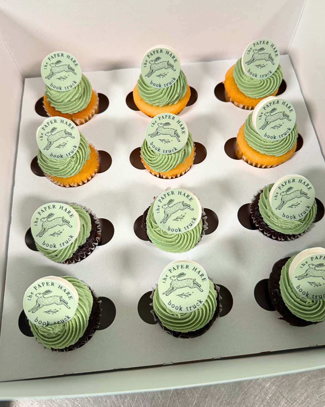 Box of cupcakes with green swirl frosting topped with circular edible toppers featuring a hare illustration and the text 'the PAPER HARE book truck'.