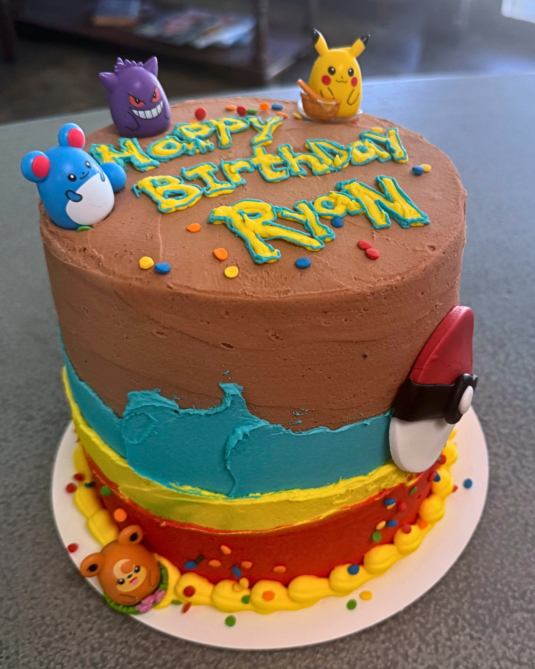 A colorful birthday cake decorated with Pokémon characters, including Gengar, Pikachu, and Rolycoly, with a rainbow-colored icing layer, confetti, and a message that says "Happy Birthday Ryan."