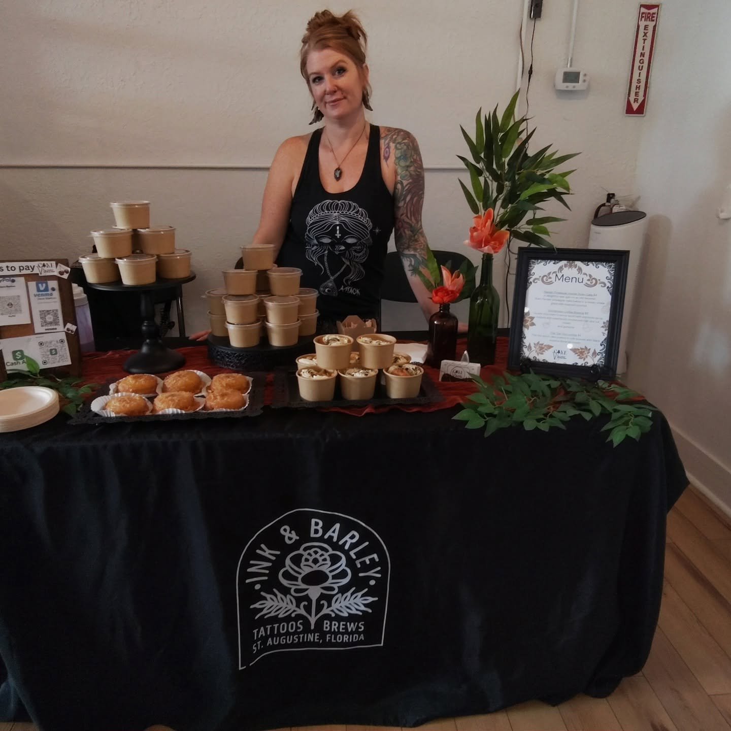 A woman with tattoos and red hair standing behind a table with small cups of food, a tray of muffins, a framed menu, and decorative plants, at a food or drink event.