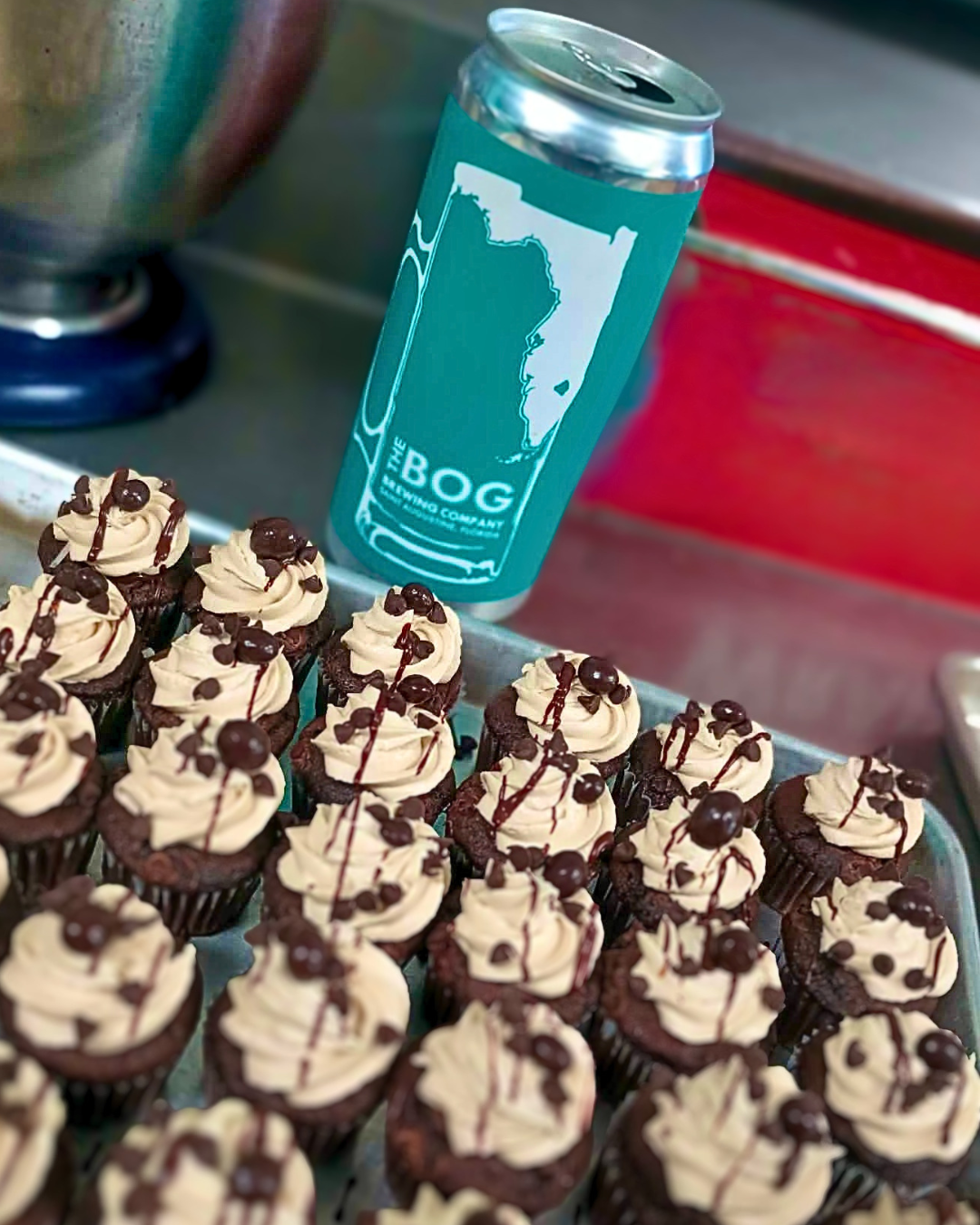 Tray of chocolate cupcakes topped with vanilla frosting and chocolate drizzles, a can of sparkling water with a map of Florida, and other drinks in the background.