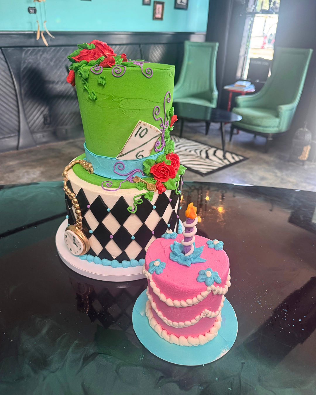 A colorful two-tier cake decorated with Alice in Wonderland theme elements, including a giant green top hat with roses and vines, a pocket watch, a playing card, and a pink cake with a candle on top with blue flowers, set on a black table in a cafe with green chairs and framed pictures on the wall in the background.