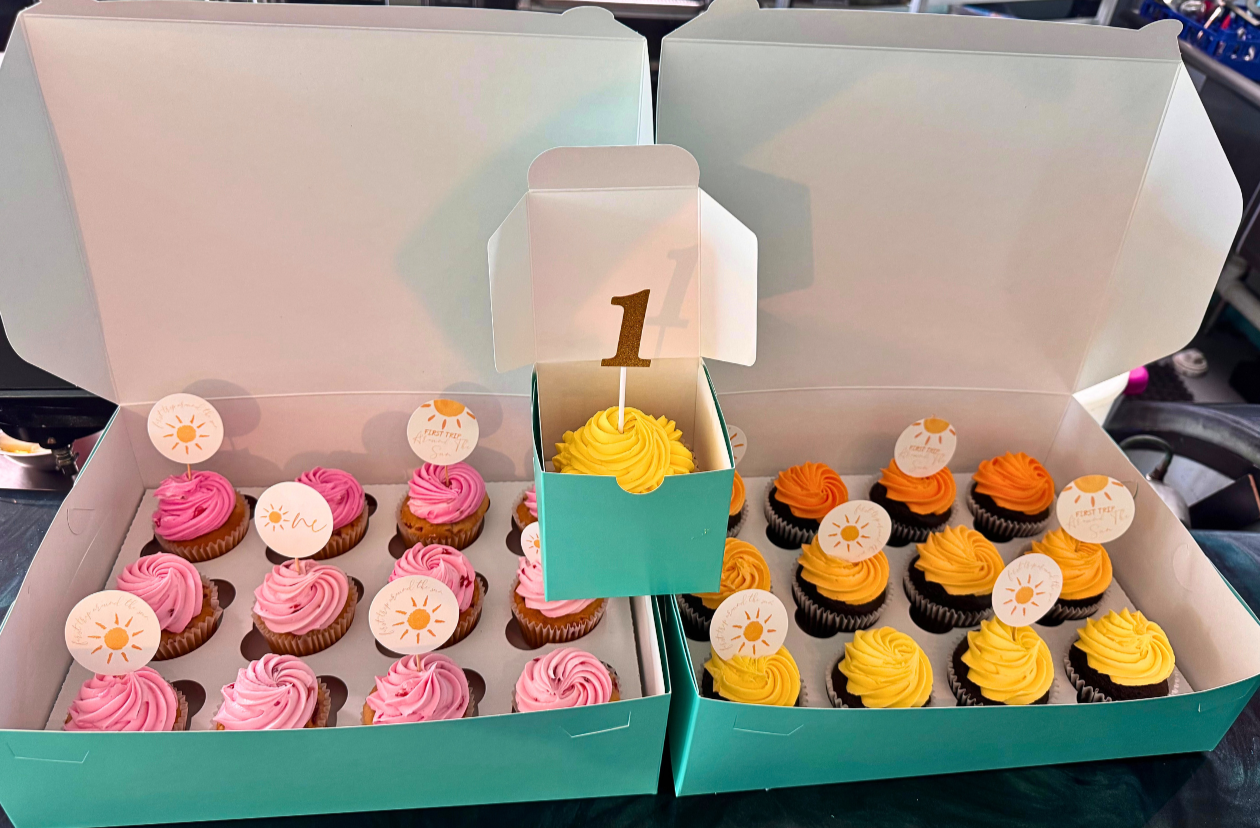 Box of pink and yellow cupcakes with decorative toppers and a cupcake with a '1' topper in a small turquoise box.