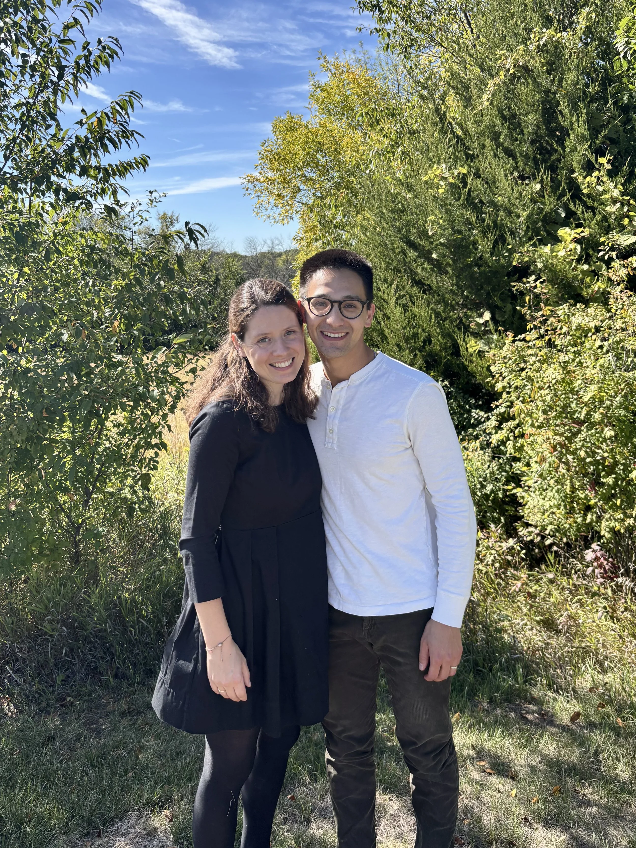 A smiling couple standing outdoors on a sunny day with trees and blue sky in the background, embracing each other.