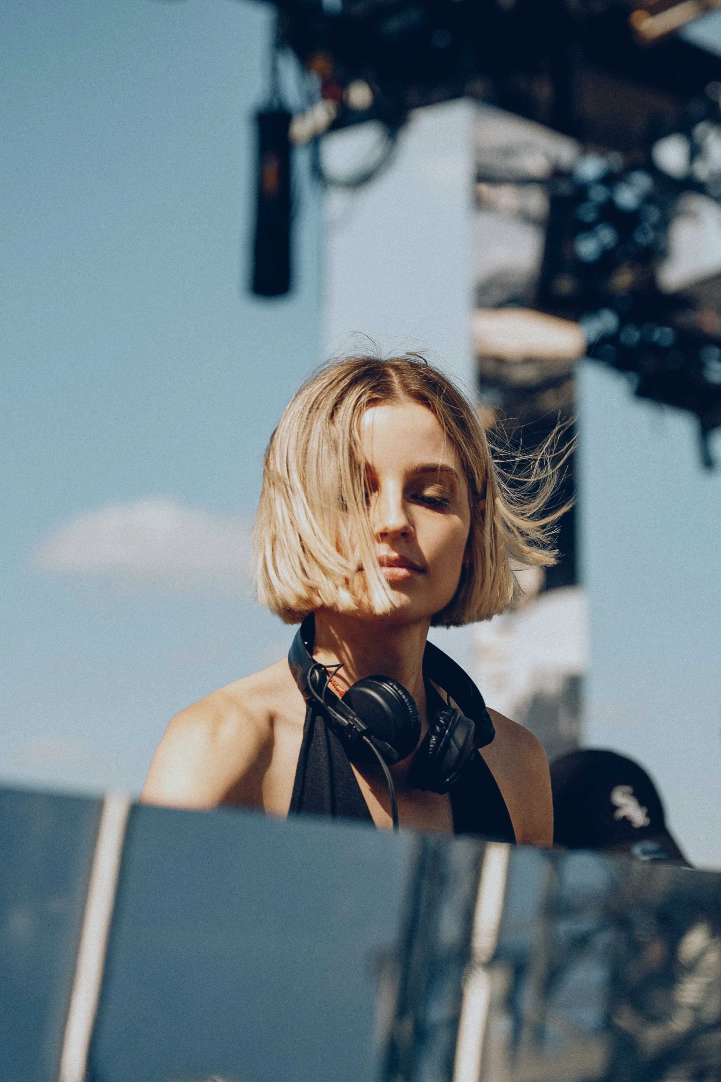 A young woman with short blonde hair wearing headphones around her neck, with her eyes closed, outdoors on a sunny day.