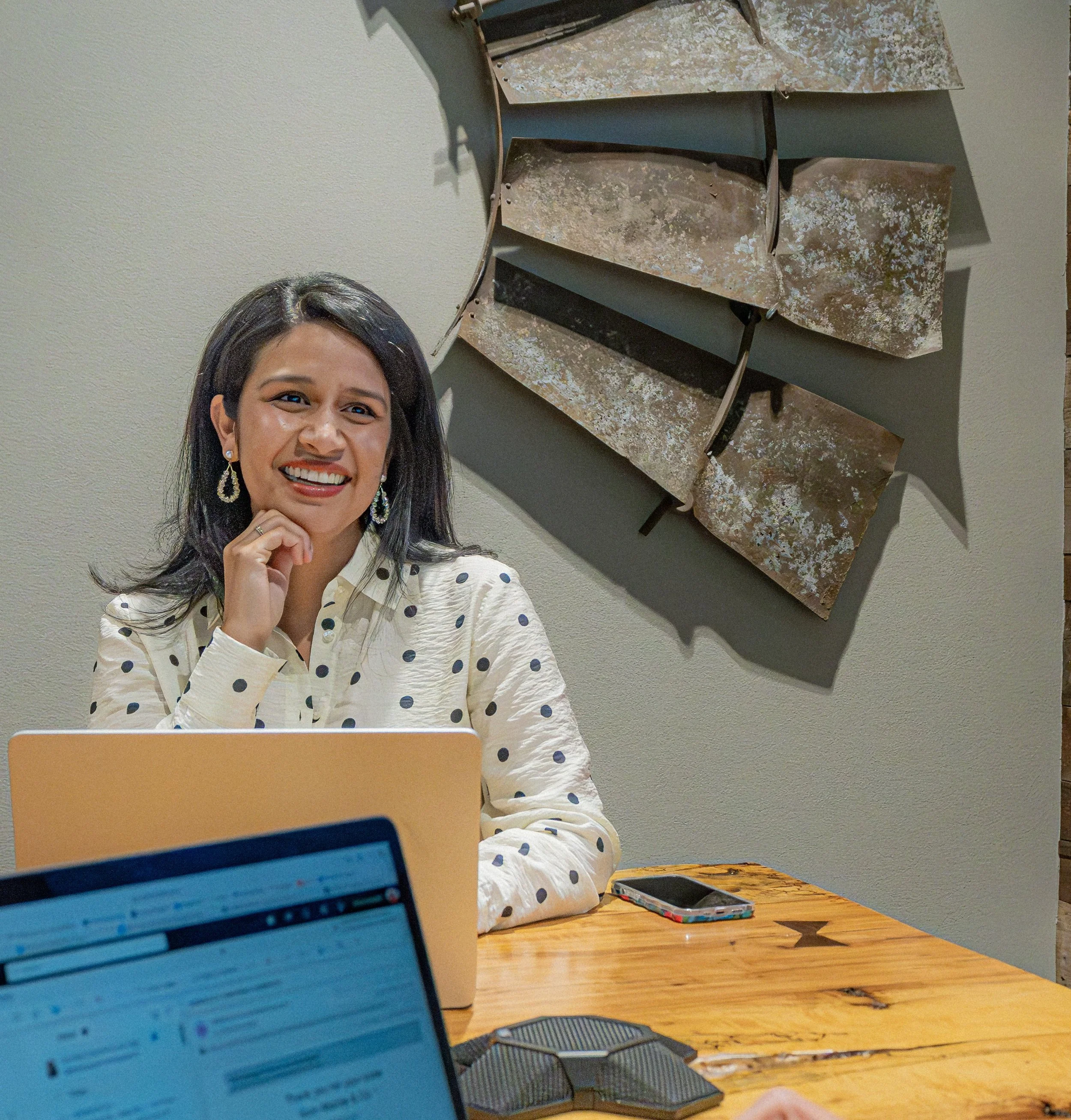 Two women seated at a wooden table with laptops and a smartphone, engaging in a discussion in an indoor setting with beige walls and decorative wall art.
