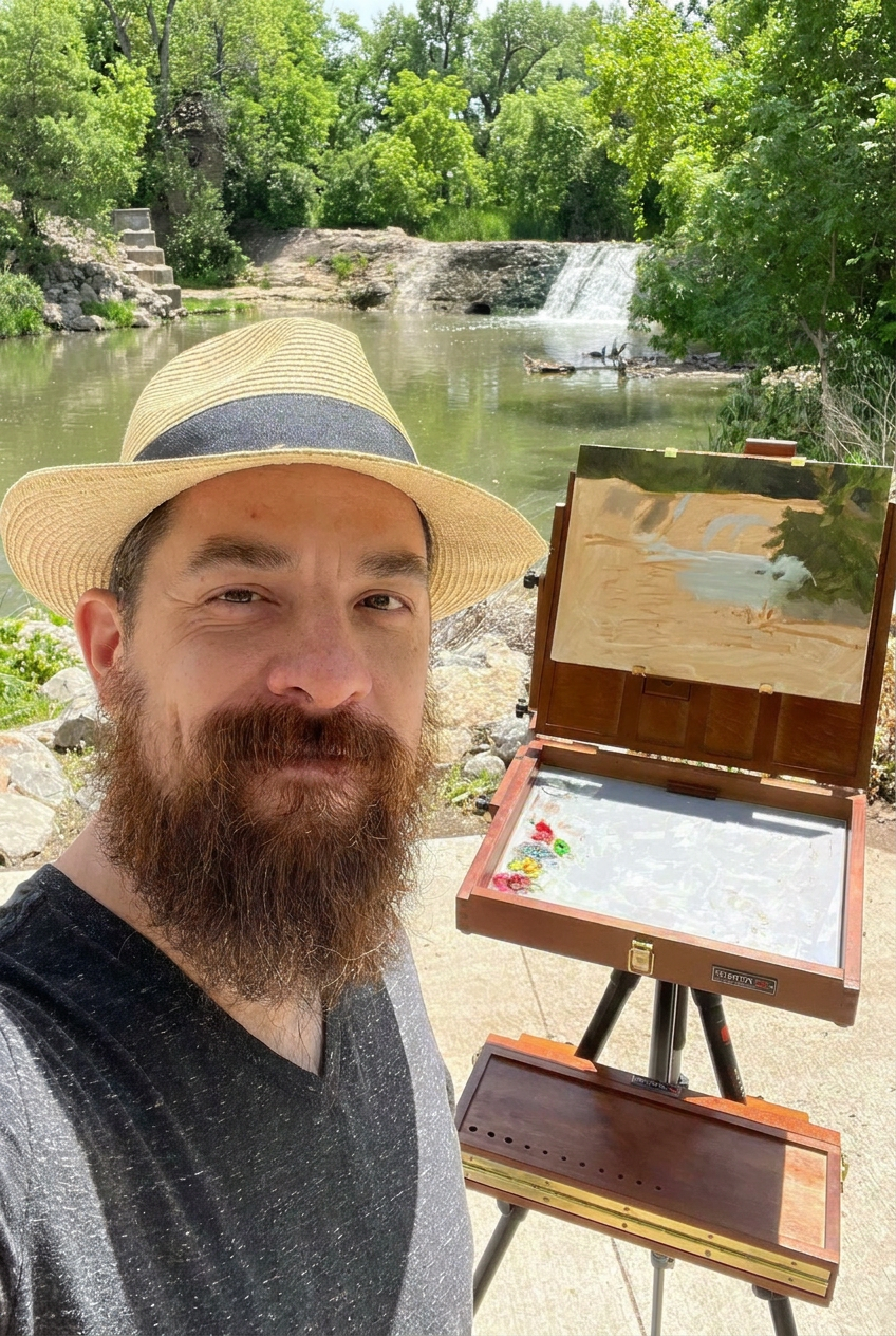 A man with a beard and a straw hat takes a selfie near a river with a small waterfall. He stands beside an open wooden art box on a tripod, which contains a palette with paints. The background features lush green trees and a peaceful outdoor setting.