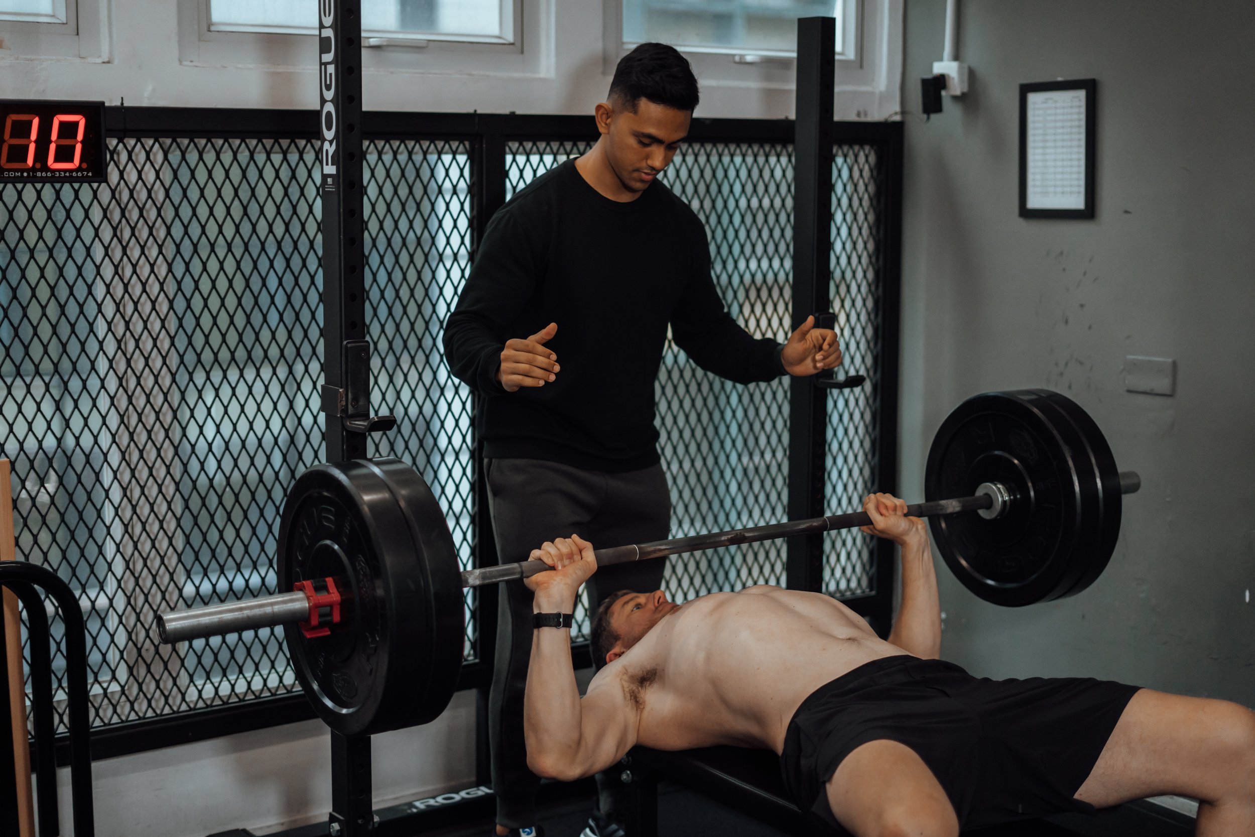 A man lying on a weight bench performing a bench press with a barbell, while another man spots him. The spotter is standing beside the bench, overseeing the exercise. The setting is an indoor gym with equipment and a digital timer visible.