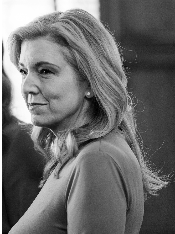 A black-and-white photo of a woman with shoulder-length wavy hair, wearing a top and stud earrings, in a side profile view, looking to the left.